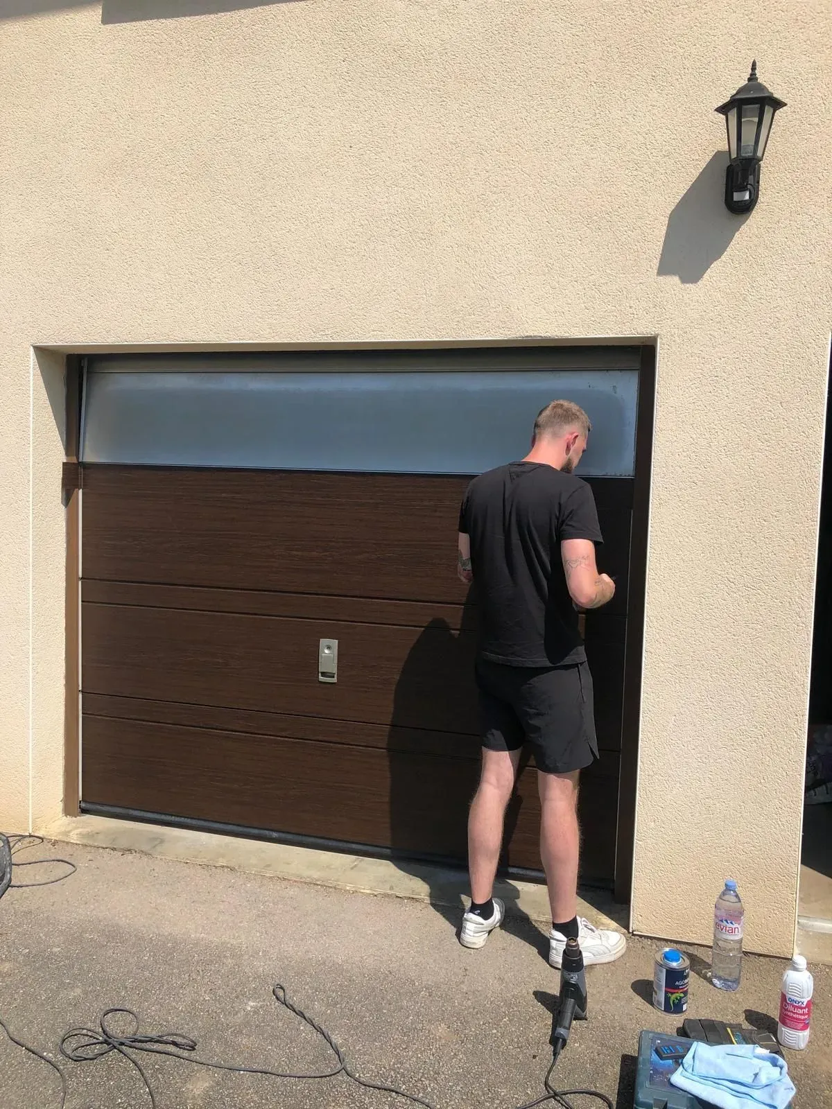 Un homme vêtu de noir installe une porte de garage à l'extérieur d'une maison beige.
