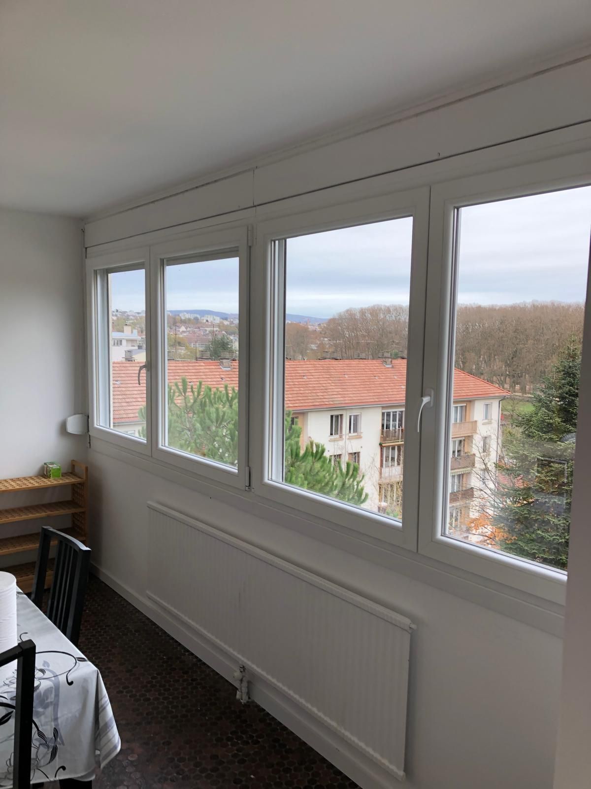 Balcon lumineux et fermé avec de grandes fenêtres, des murs blancs, un radiateur et une vue sur les toits et les arbres.