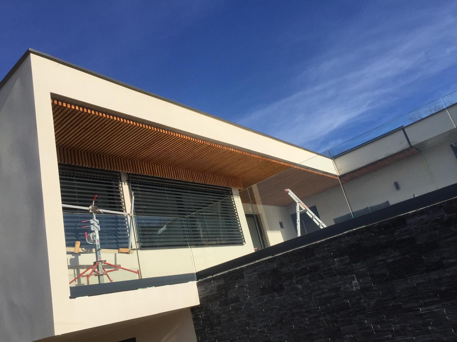 Extérieur d'une maison moderne avec balcon, auvent en bois à claire-voie et mur noir sous un ciel bleu.