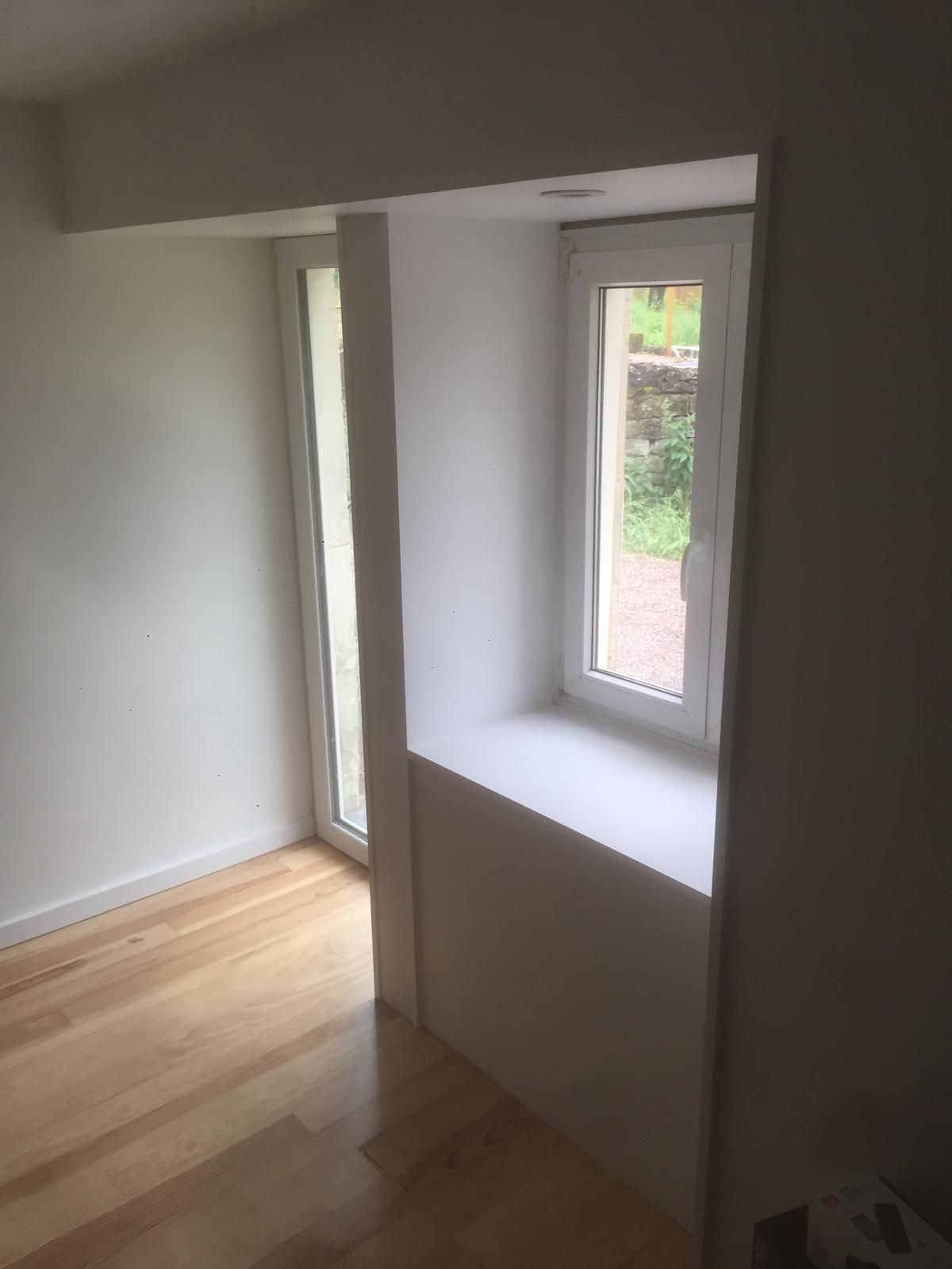 Pièce vide avec parquet clair, murs blancs et deux fenêtres étroites laissant entrer la lumière du jour.