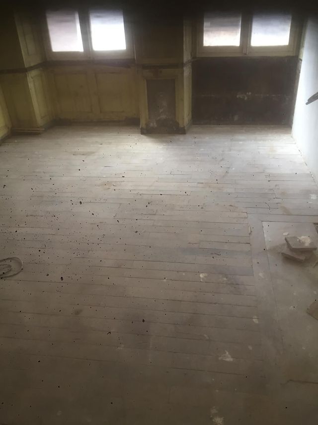Pièce en bois vide, au parquet usé, avec deux fenêtres laissant entrer une faible lumière.