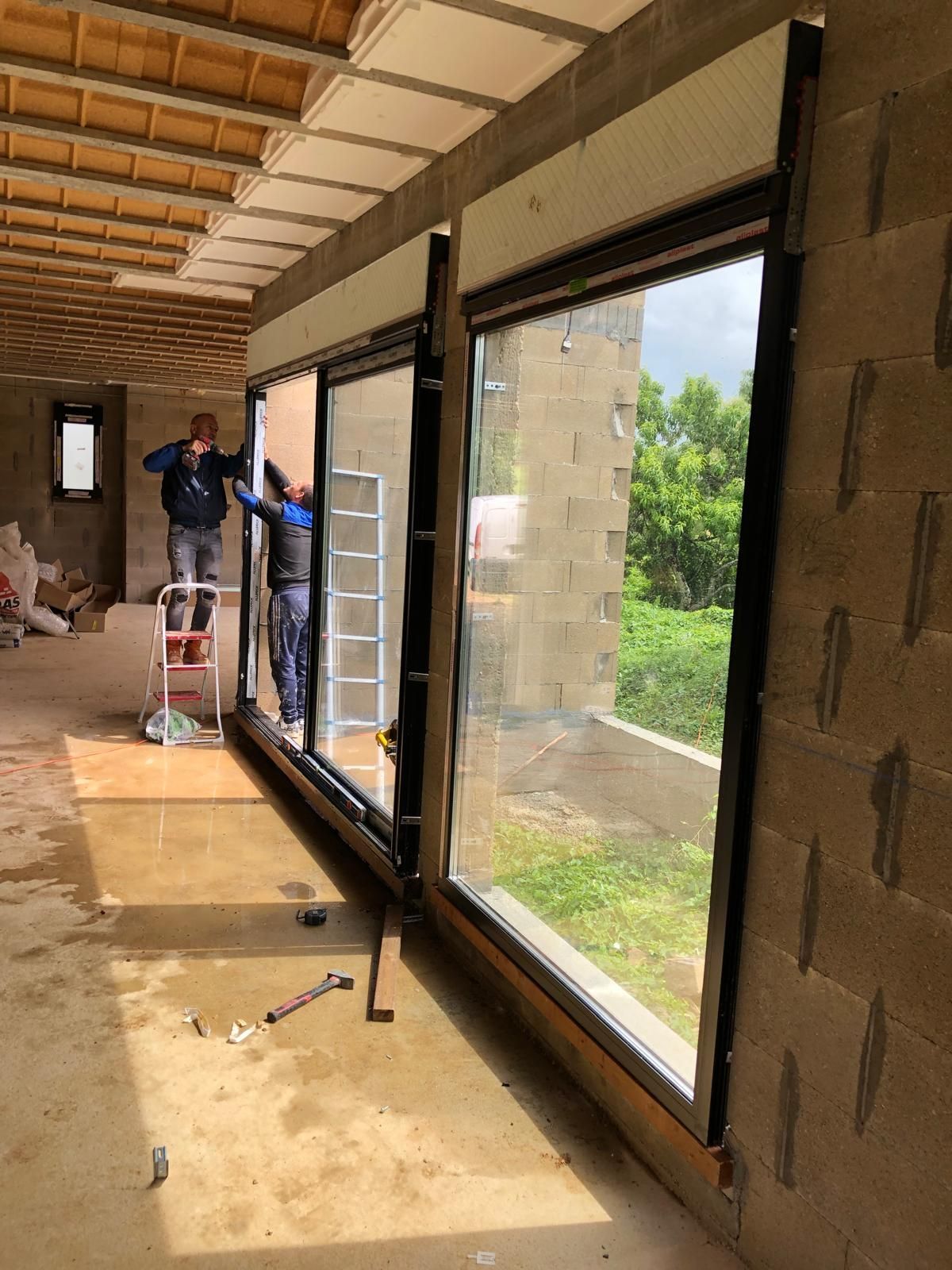 Des ouvriers installent de grandes fenêtres dans une maison inachevée aux murs apparents.