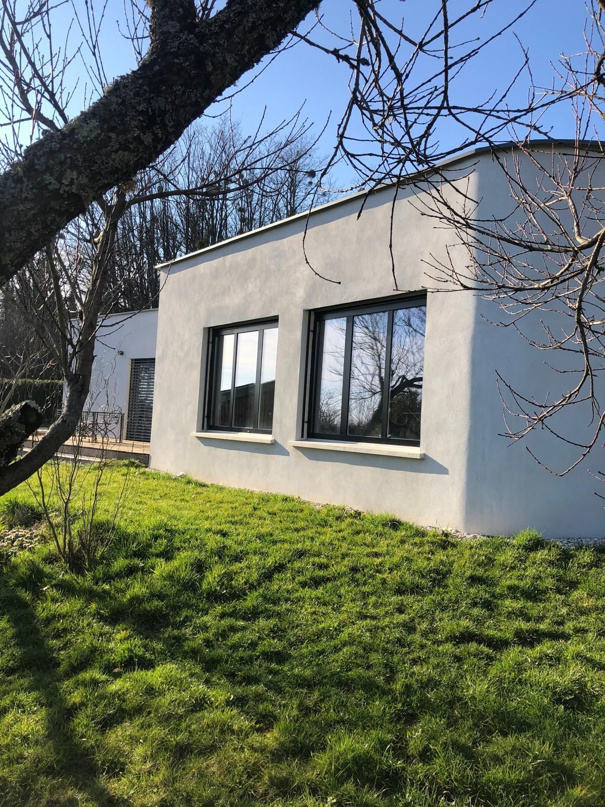 Maison blanche moderne aux fenêtres à cadre sombre, située sur un versant herbeux ensoleillé, aperçue à travers les branches des arbres.