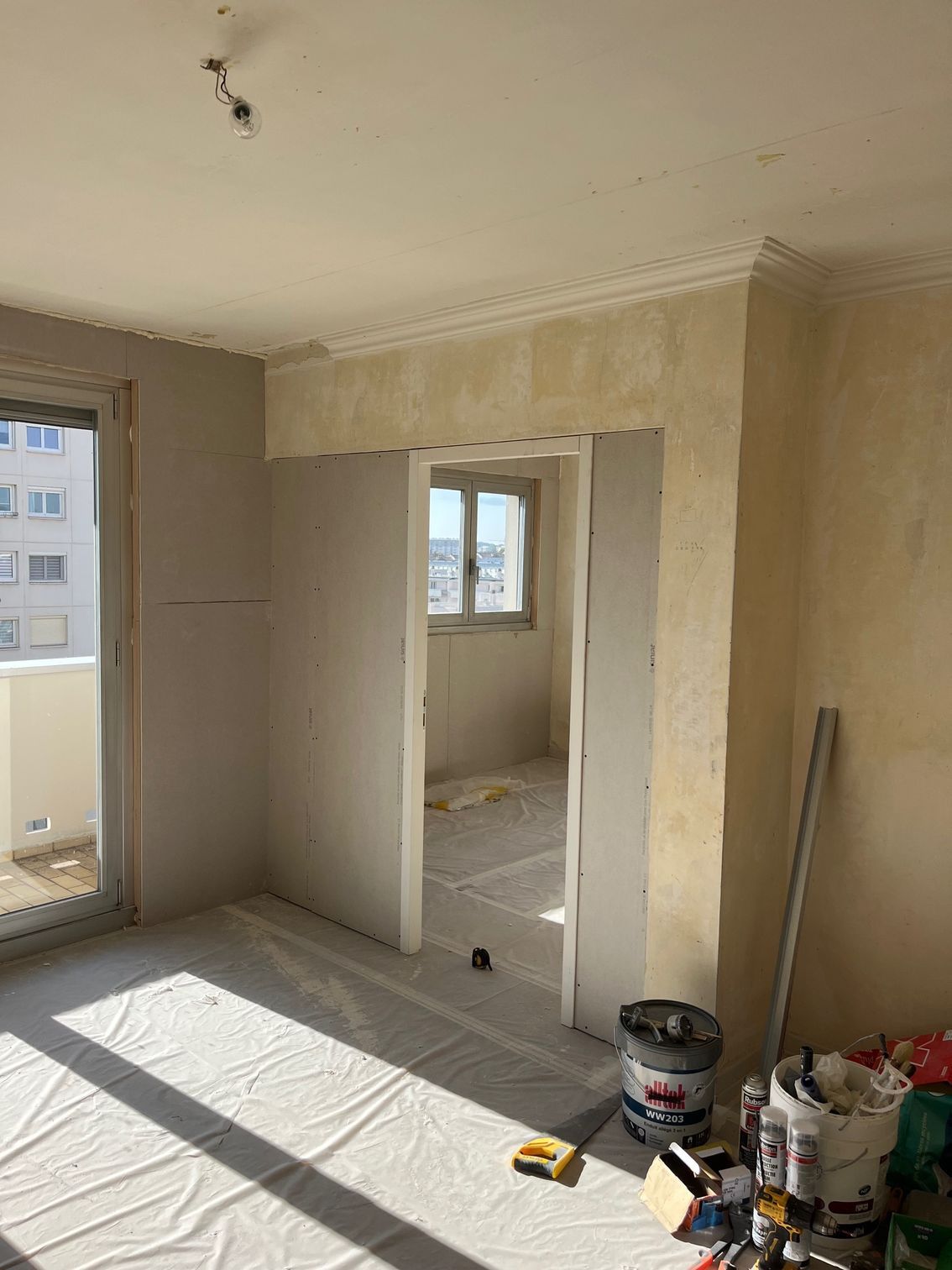 Pièce d'appartement inachevée avec des murs beiges, de la lumière naturelle, une porte ouverte et des matériaux de rénovation au sol.