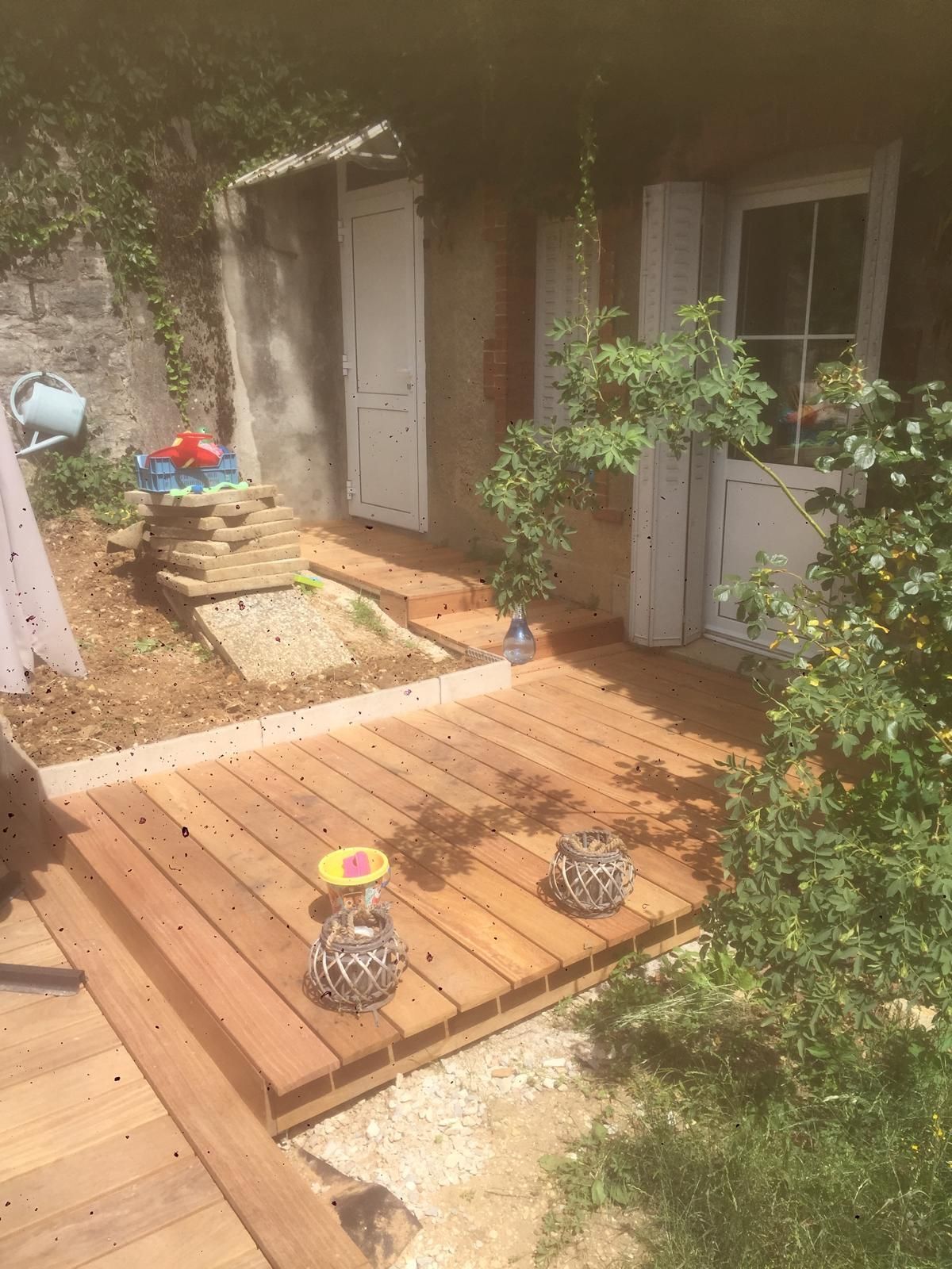 Petit patio de jardin avec terrasse en bois, plantes en pot et une porte blanche à côté d'un mur de briques.