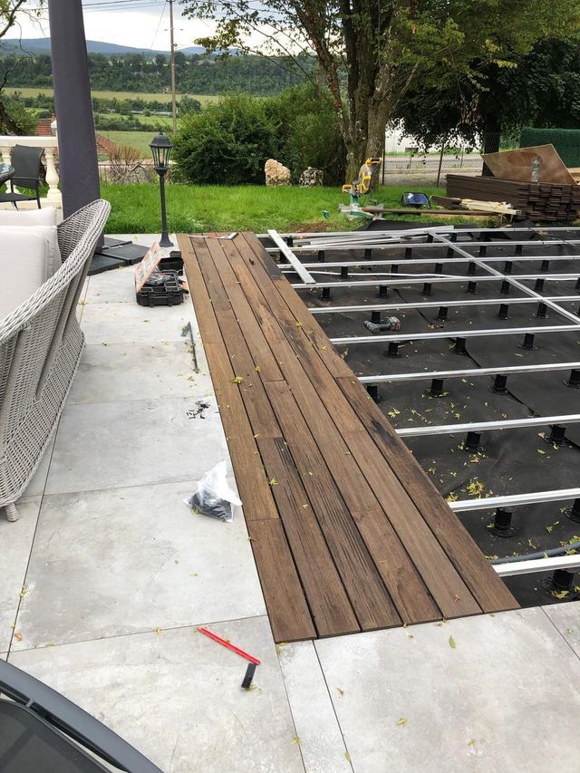 Terrasse en bois partiellement construite, structure apparente et dalles de béton à côté d'une maison.