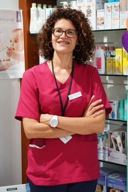 Una mujer con una blusa rosa está parada con los brazos cruzados en una farmacia.