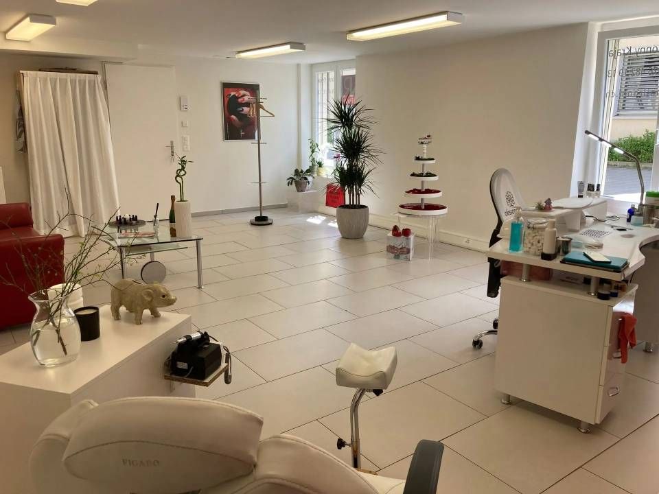 Innenansicht eines Nagelstudios mit weißen Möbeln, Pflanzen und einem Wartebereich. Hell erleuchtet durch große Fenster.