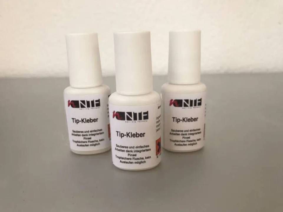 Drei weiße Flaschen „Tip-Kleber“ Nagelkleber mit roter und schwarzer Schrift auf hellgrauem Grund.