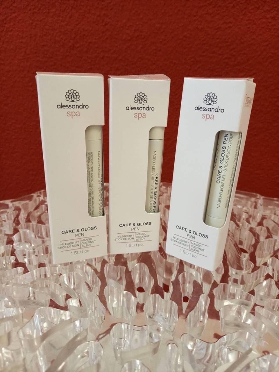 Drei weiße Schachteln mit Lippenpflegeprodukten von Alessandro auf einer Glasoberfläche vor rotem Hintergrund.