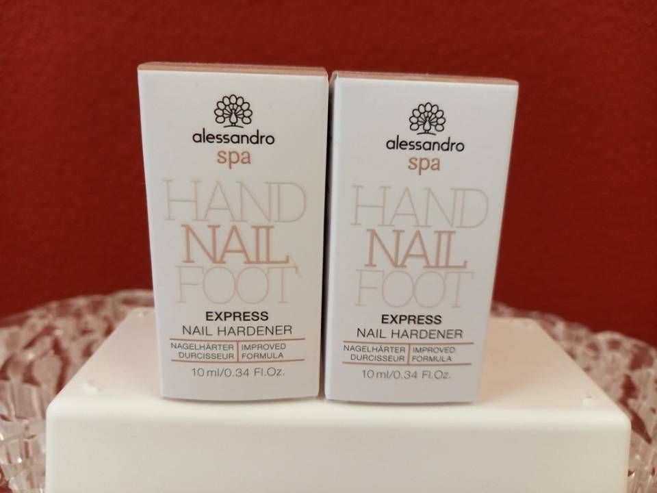 Zwei weiße Alessandro Spa Nagelhärter-Dosen mit goldener Schrift auf weißem Grund vor rotem Hintergrund.