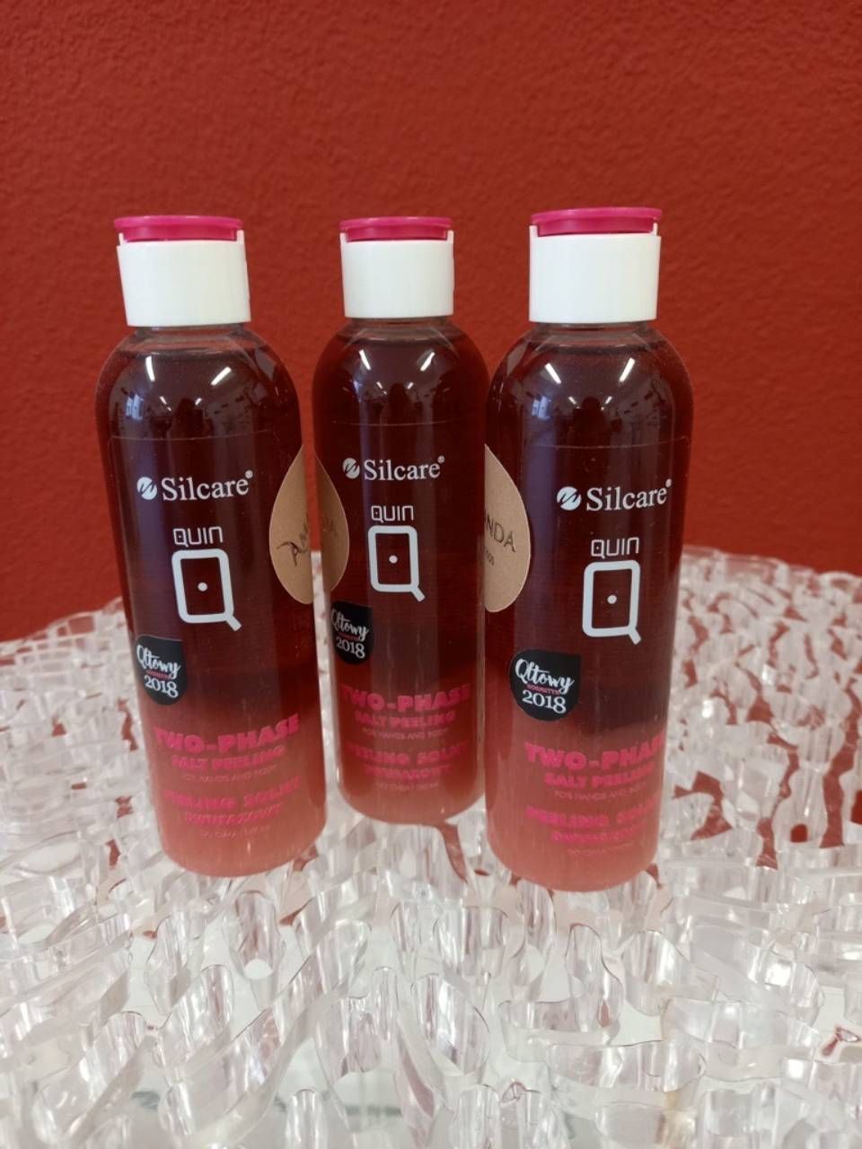 Drei Silcare-Nagelhautöl-Flaschen auf strukturierter Fläche vor rotem Hintergrund, rotbraun mit weißen und rosa Deckeln.