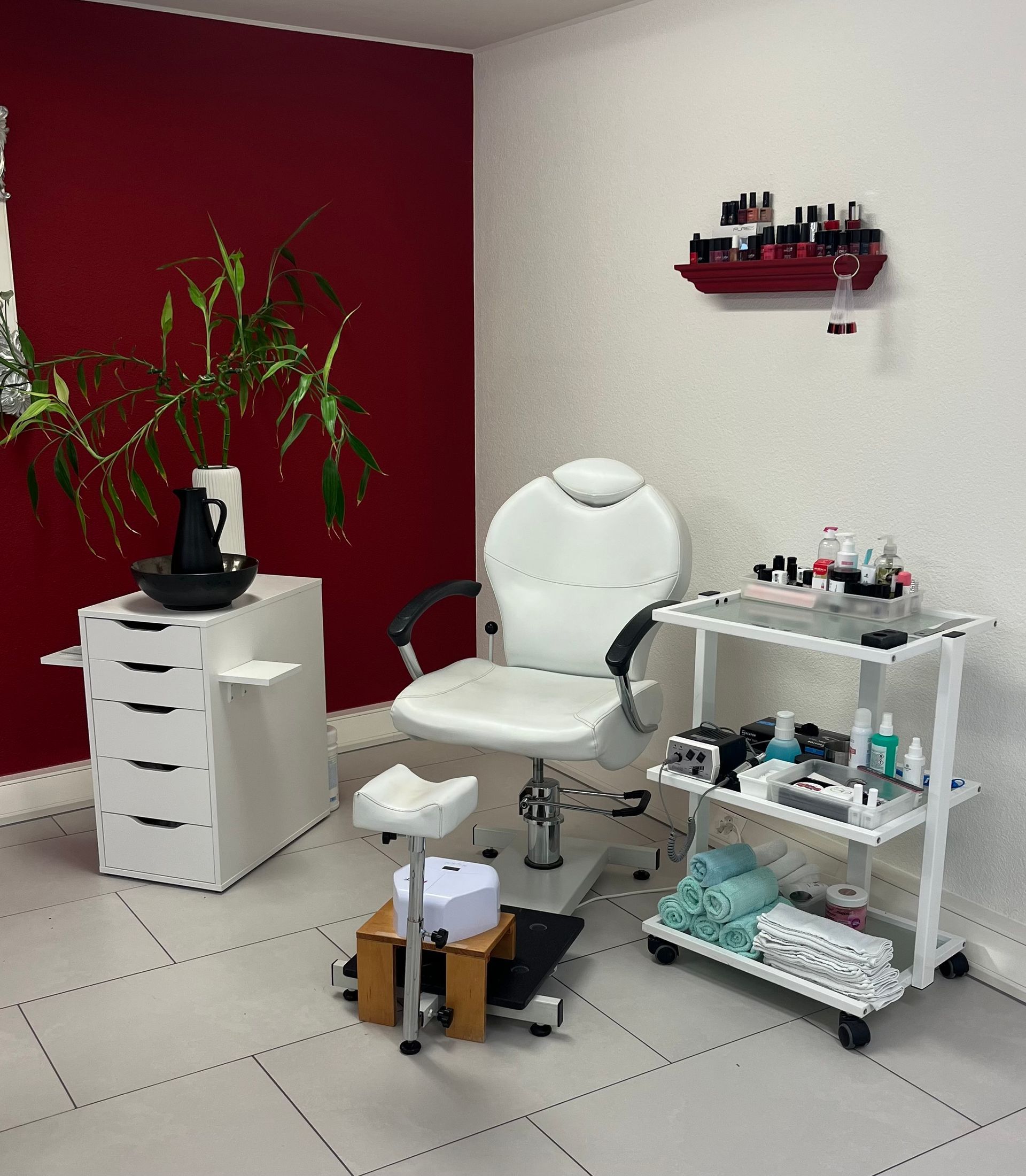 Innenansicht eines Nagelstudios mit weißem Stuhl, Hocker und Wagen auf gefliestem Boden vor rot-weißen Wänden.