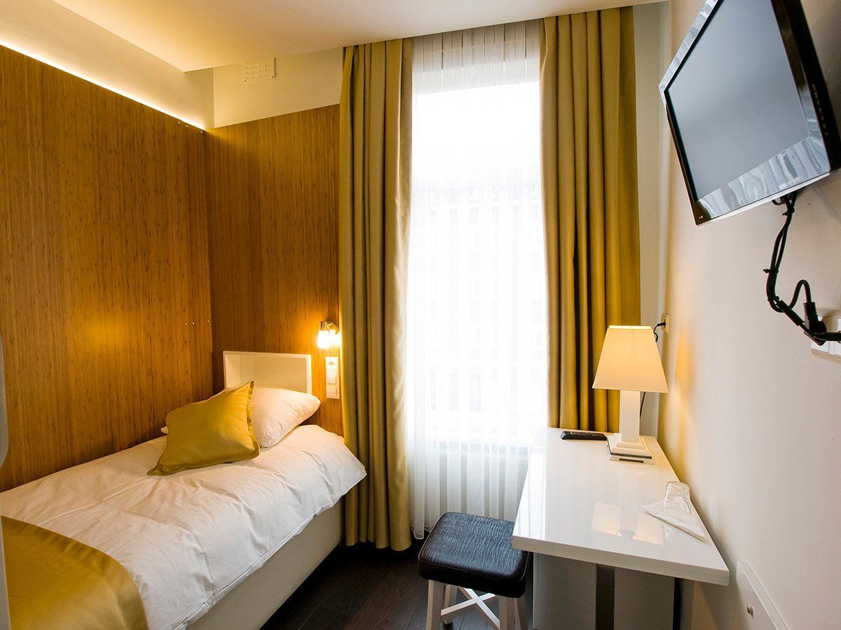 Small hotel room with a single bed, desk, wall-mounted TV, and curtained window.