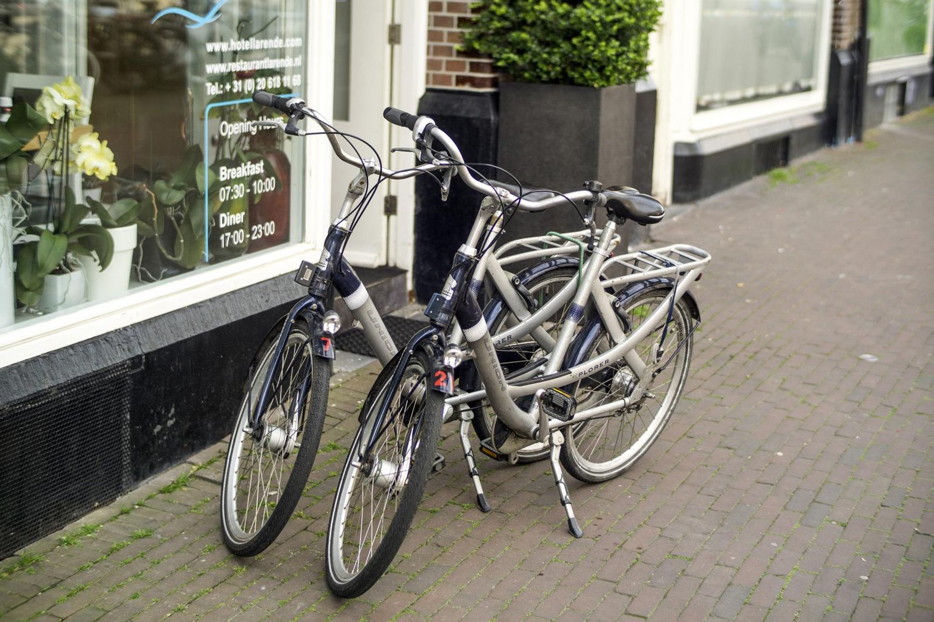 Two bicycles parked on a sidewalk beside a storefront window and brick wall.