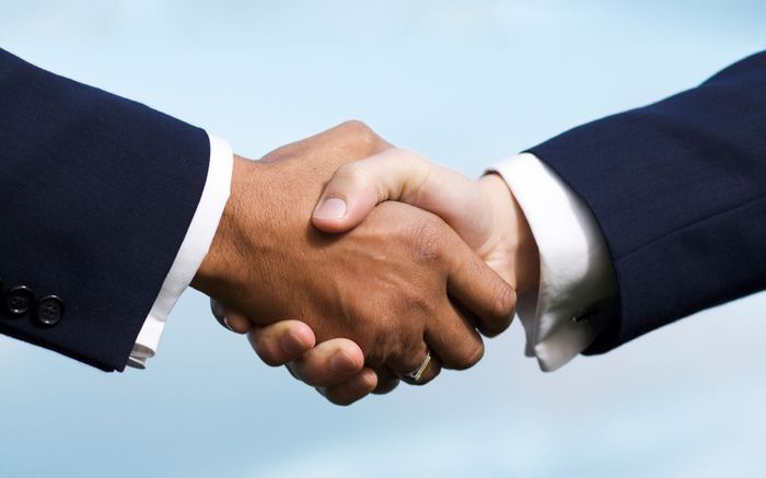 Two people in dark suits shaking hands against a blue sky background