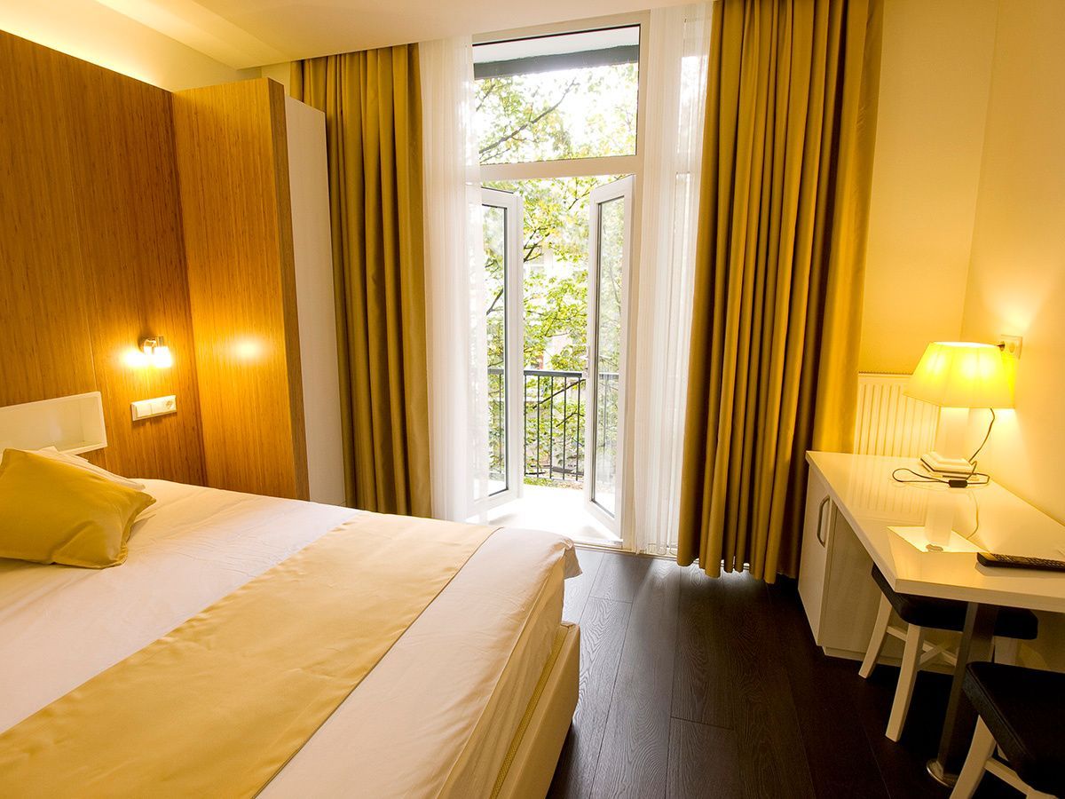 Bright hotel room with a bed, warm lamps, desk, and balcony doors with curtains.