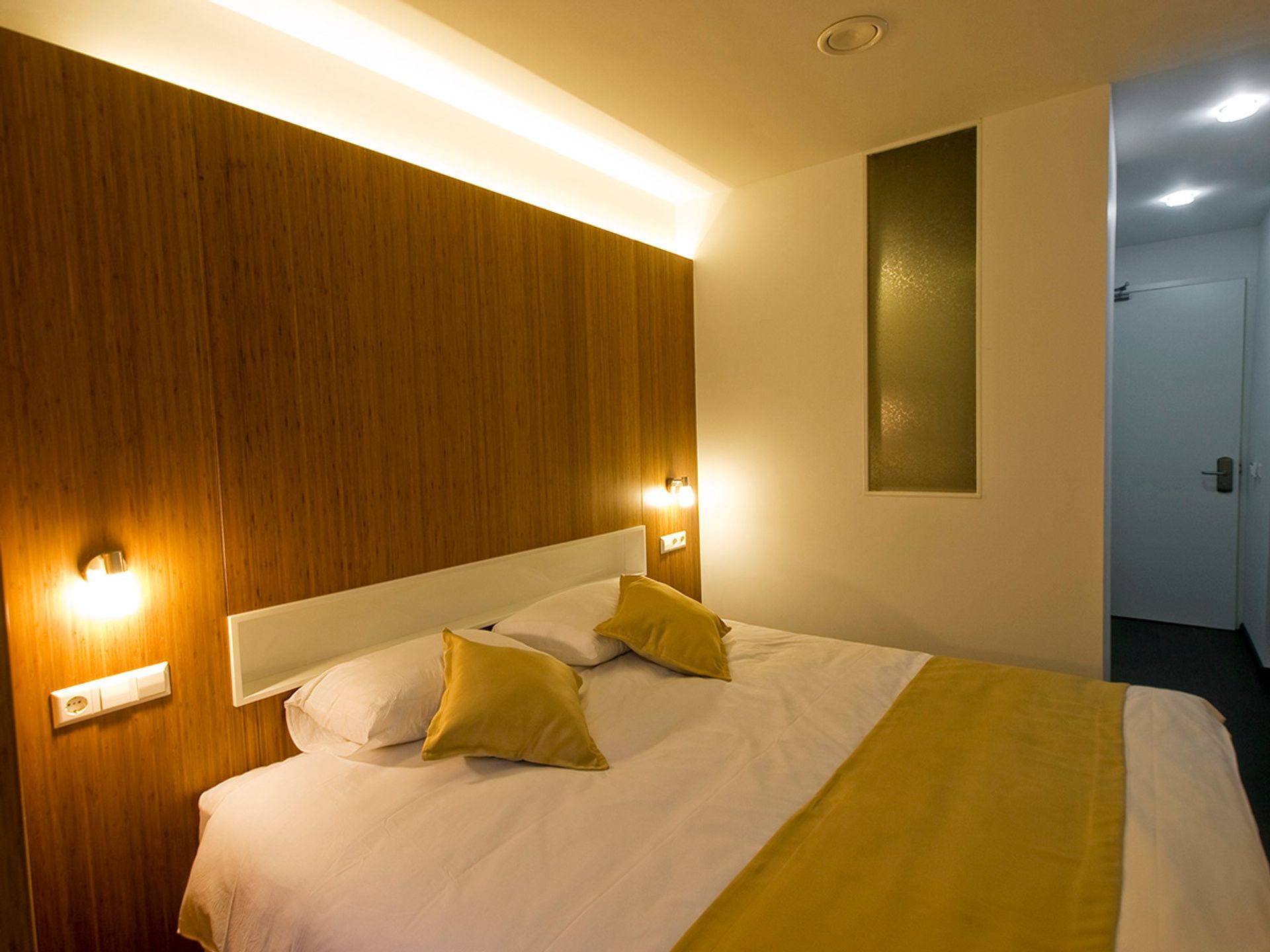 Modern hotel bedroom with double bed, yellow accents, wood-paneled wall, and soft lighting