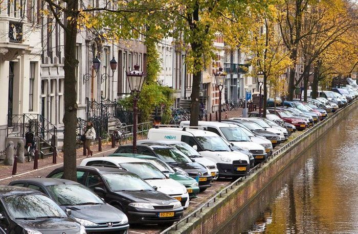 Parked cars line a canal beside historic buildings and autumn trees in a European street.