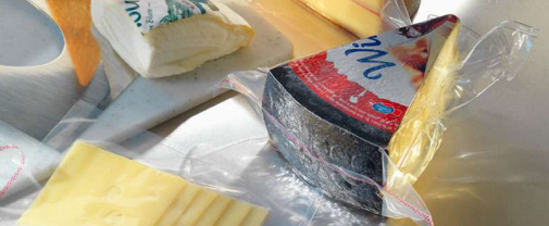 Verschiedene verpackte Lebensmittel auf einem Tisch, darunter Brot, Butter und ein runder Käselaib in Plastikfolie.