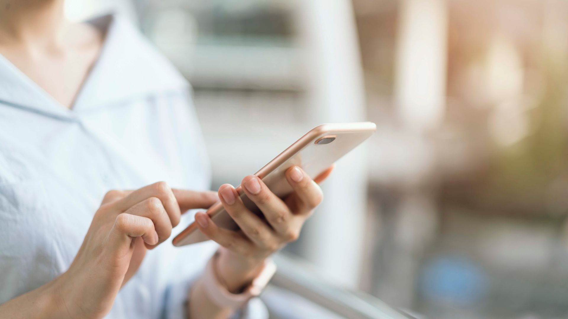 Person, die in der Nähe eines Fensters ein Smartphone benutzt, im Hintergrund ist ein verschwommener Büroraum zu sehen.