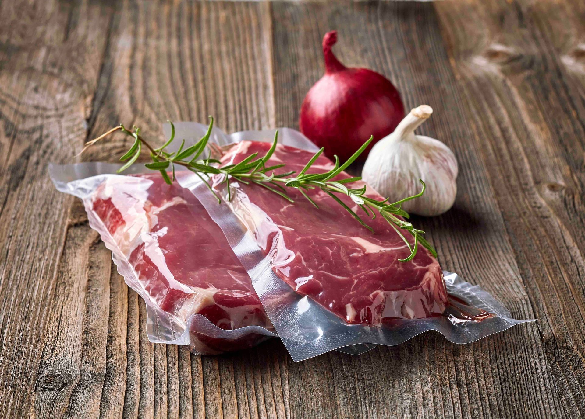 Rohe Steaks mit Rosmarin, roten Zwiebeln und Knoblauch auf einem Holztisch