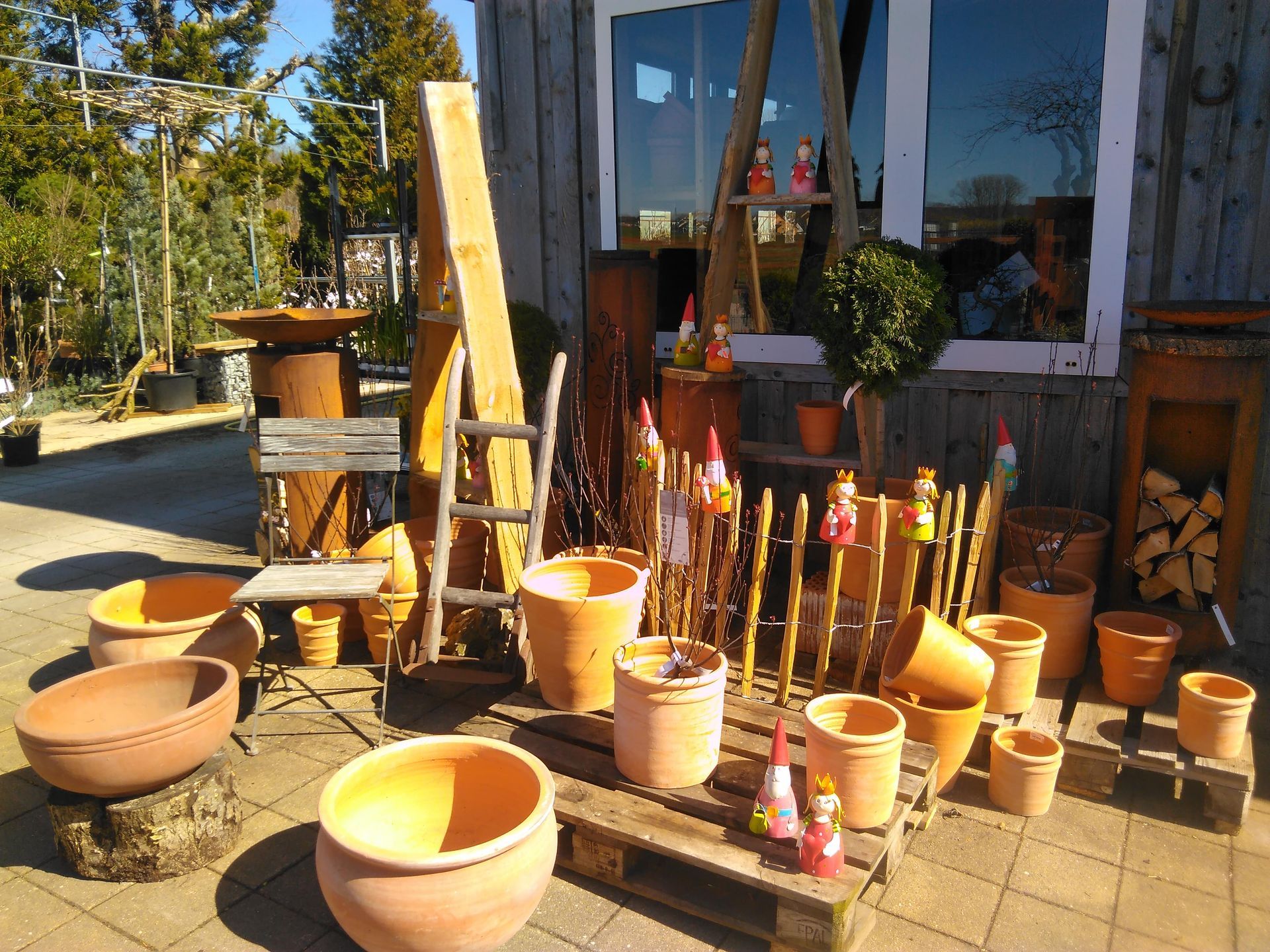 Außenausstellung von Terrakottatöpfen, Holzleitern und Gartenornamenten vor einem Holzgebäude.