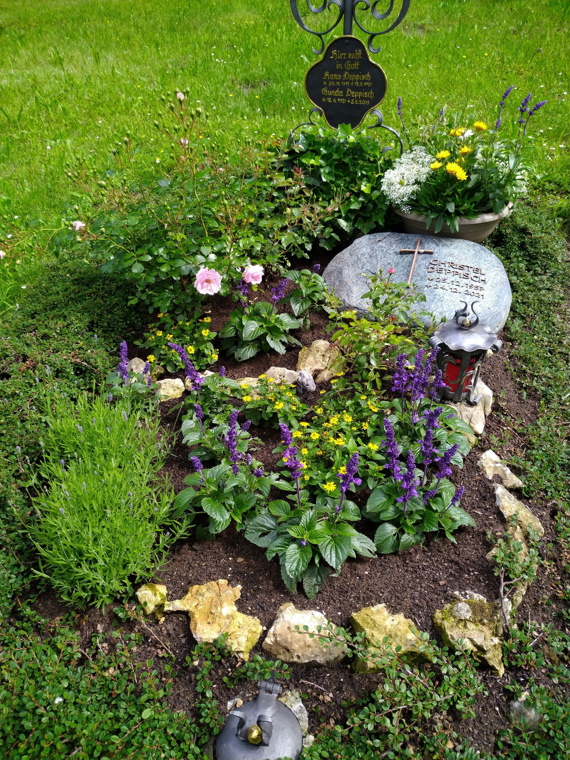 Ein farbenfrohes Grab mit Blumen, Pflanzen, Steinen und einem Grabstein auf einer Rasenfläche.