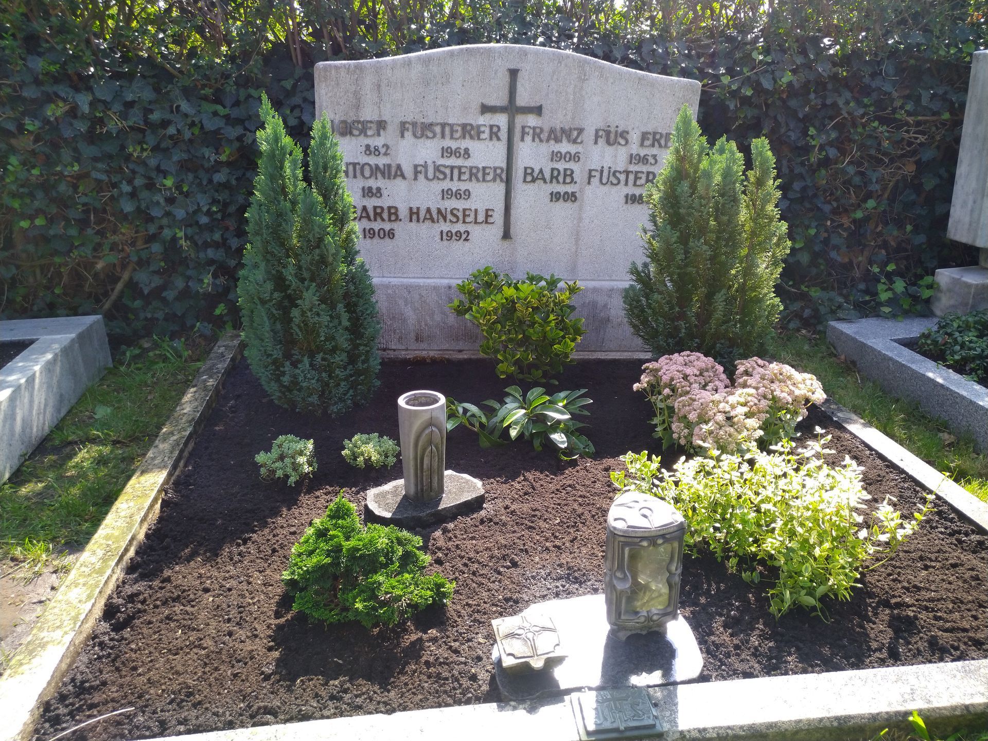 Grabstein mit Namen, Daten und einem Kreuz, umgeben von Grün und Blumen auf einem Friedhof.