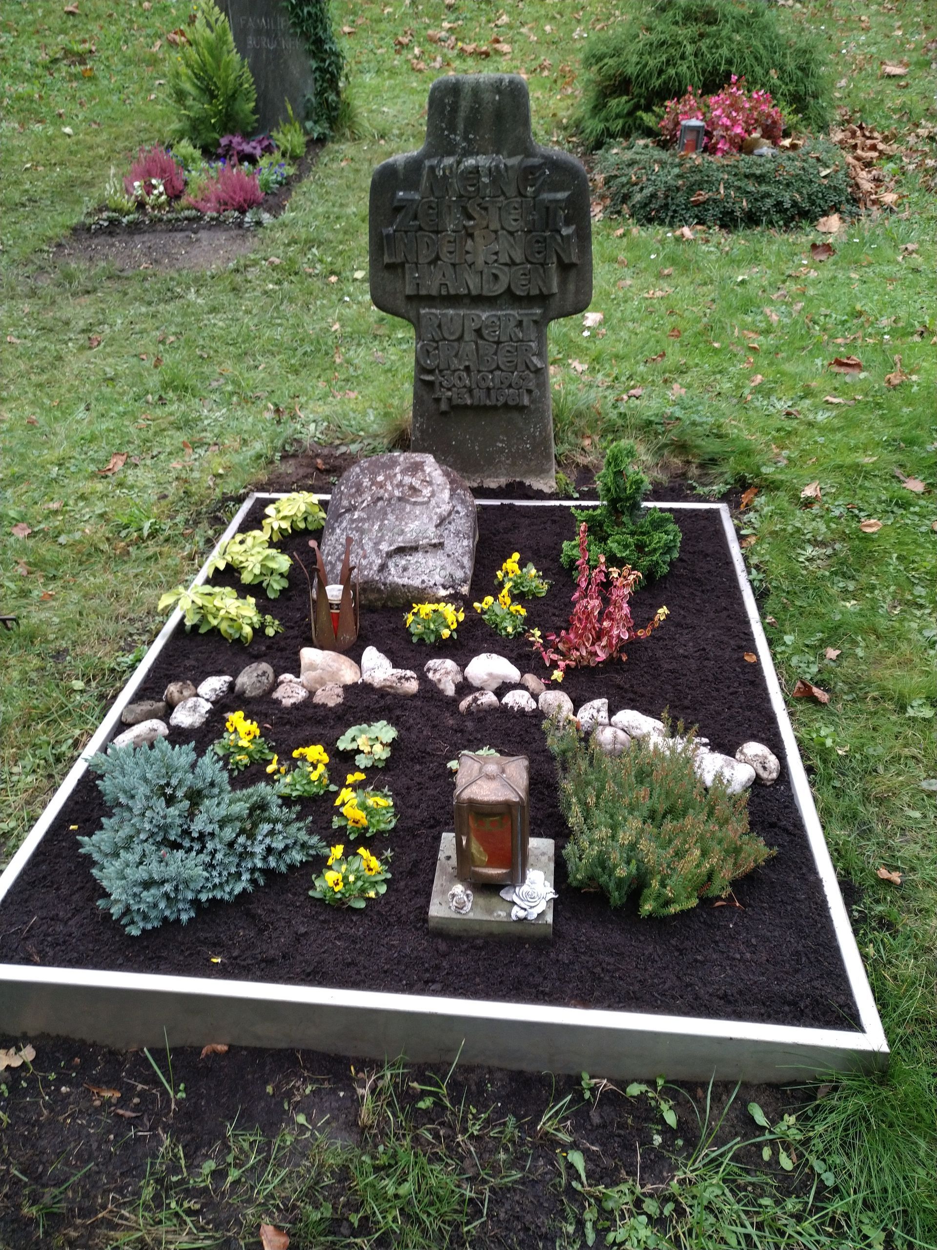 Grab mit Steinkreuz, dunkler Erde und bunten Pflanzen. Ein heller Rahmen umschließt das Grab.