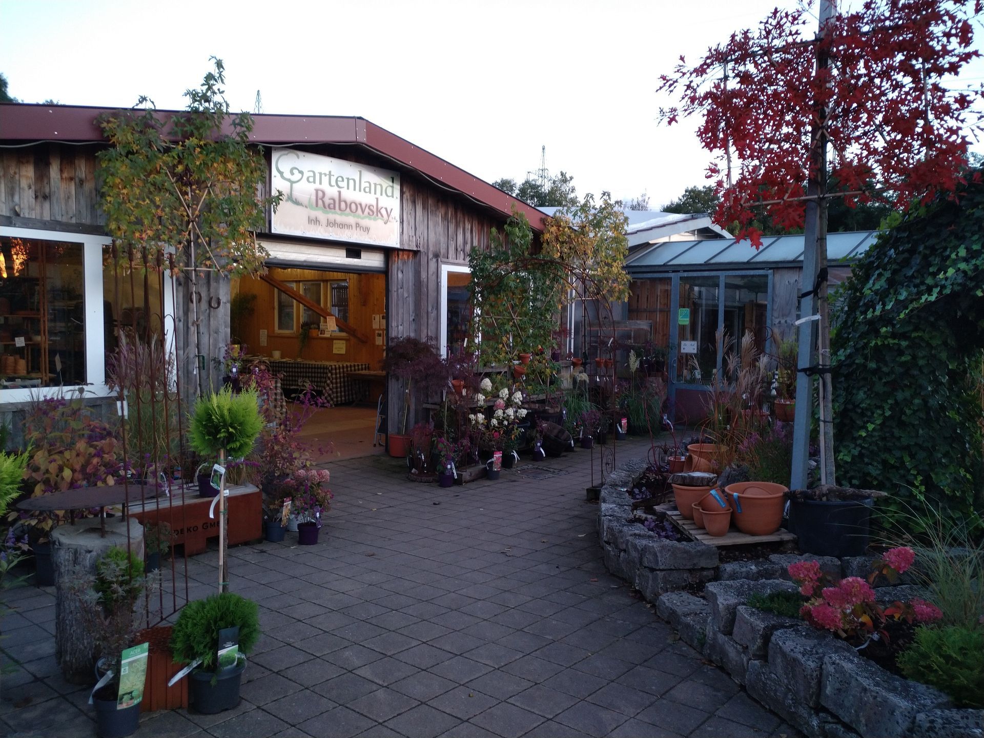 Eingang zum Gartencenter mit holzgetäfeltem Gebäude, Pflanzen und Ziegelweg.