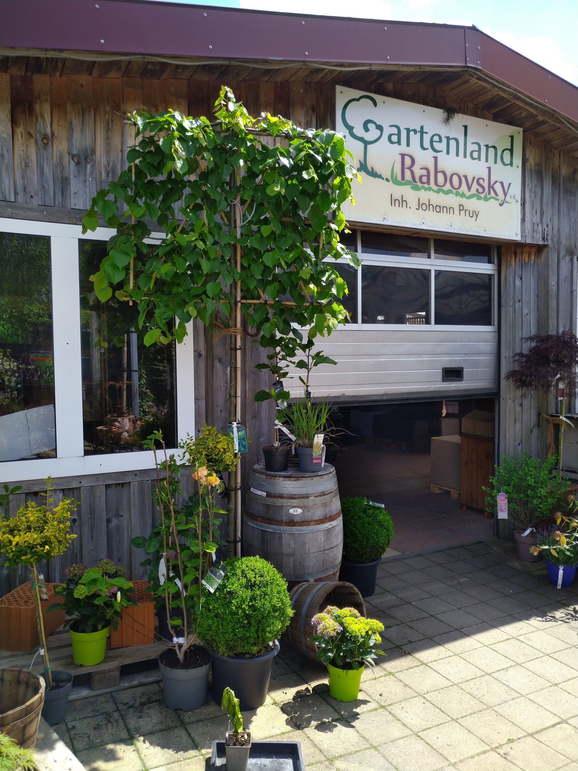 Gartenland Babovsky Gartenshop mit Pflanzen, Fässern und offenem Garagentor.