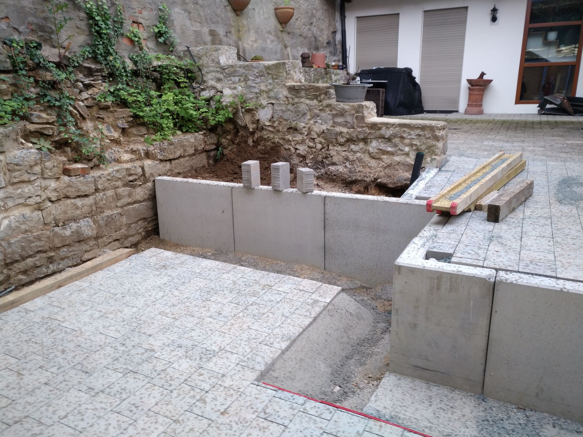 Bauarbeiten im Hinterhof: Steinstufen, Stützmauern, graue Pflastersteine, Baumaterialien und Pflanzen.