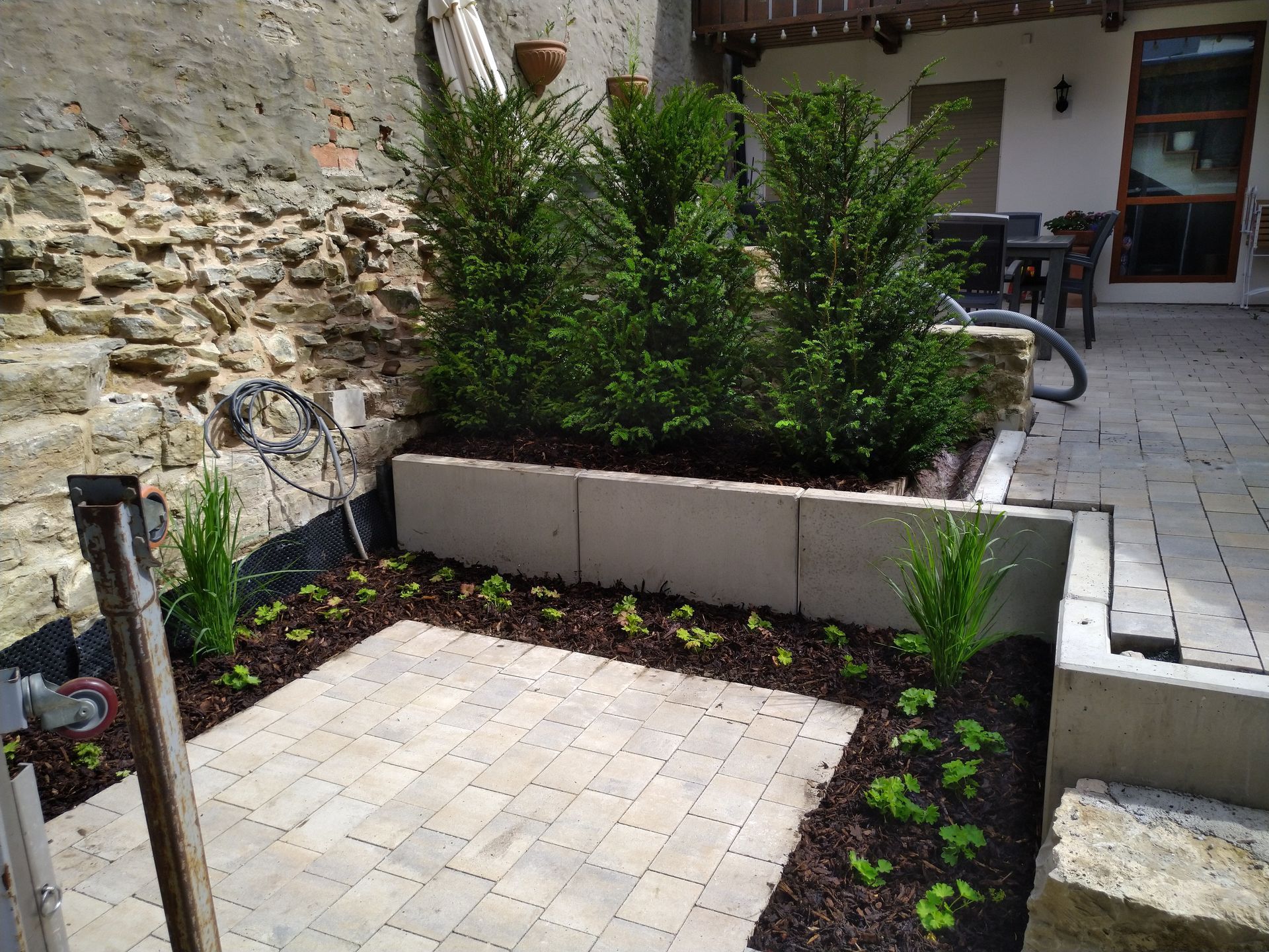 Steinterrasse mit Hochbeet und üppigen grünen Pflanzen vor einer Steinmauer.