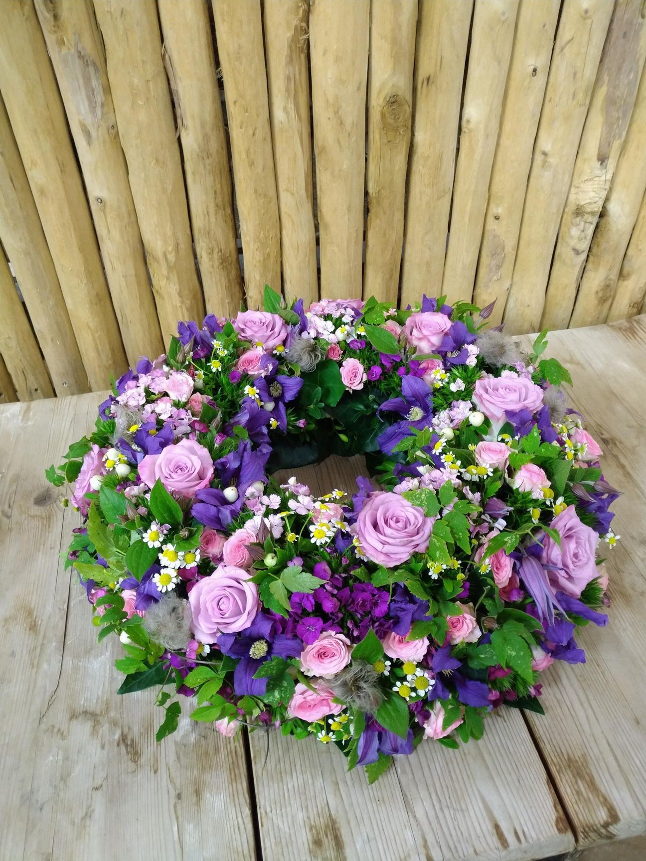 Trauerkranz mit lila und rosa Rosen, Grünpflanzen und lila Blumen auf einem Holztisch.