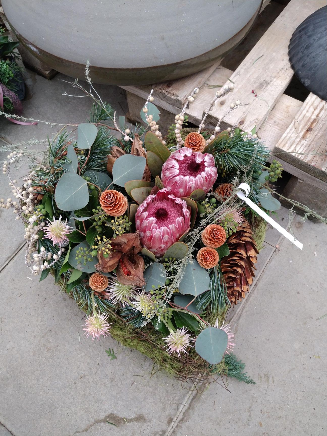 Herzförmiges Blumenarrangement mit rosa Protea, Tannenzapfen, Grünpflanzen und Moos auf einer Holzoberfläche.
