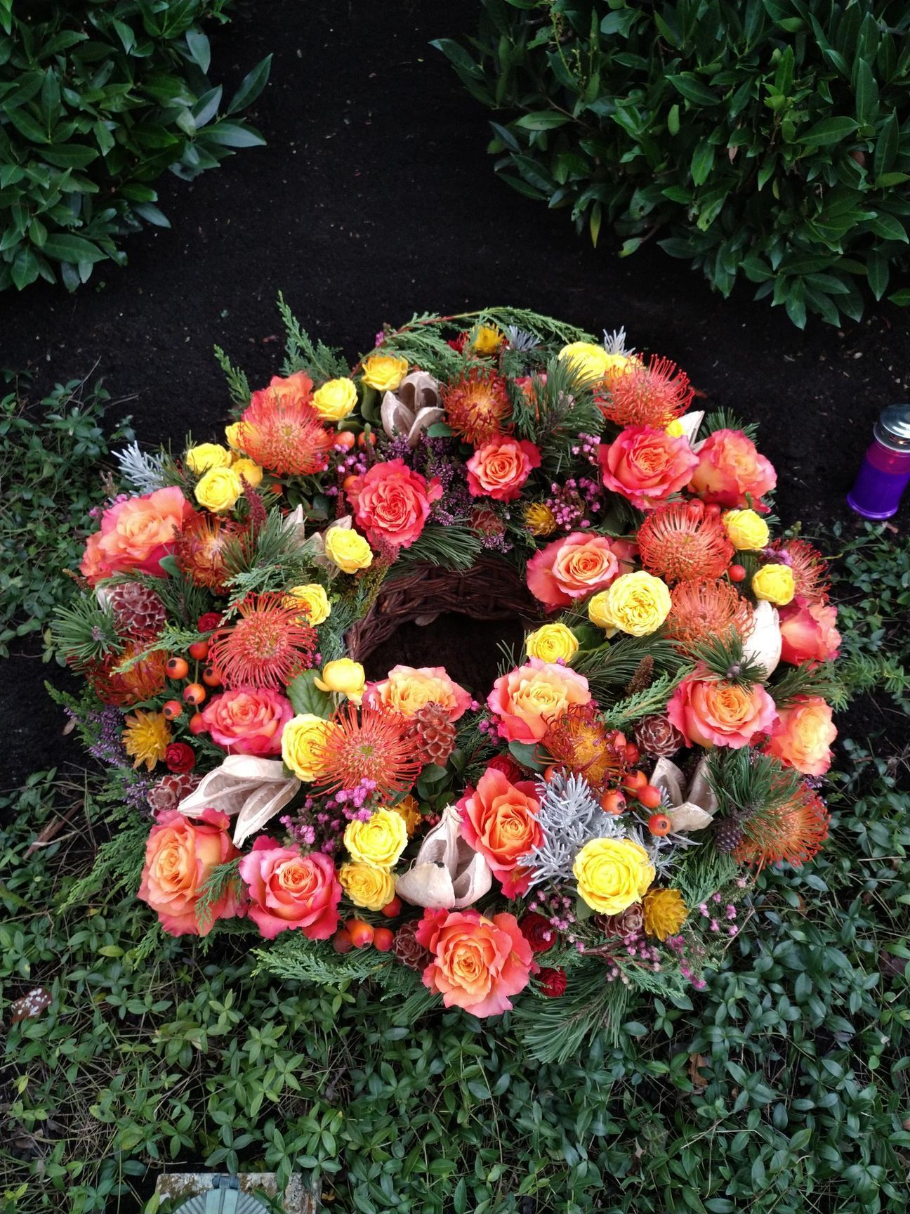 Kranz aus orangefarbenen, gelben und roten Blumen auf grünem Laub, wahrscheinlich an einer Grabstätte.