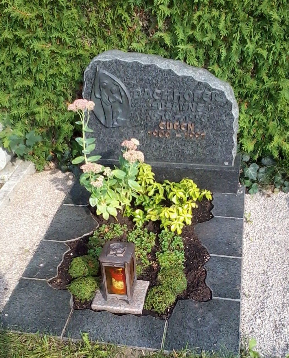 Grabstein mit dunklem Stein, Pflanzen und einer Laterne.