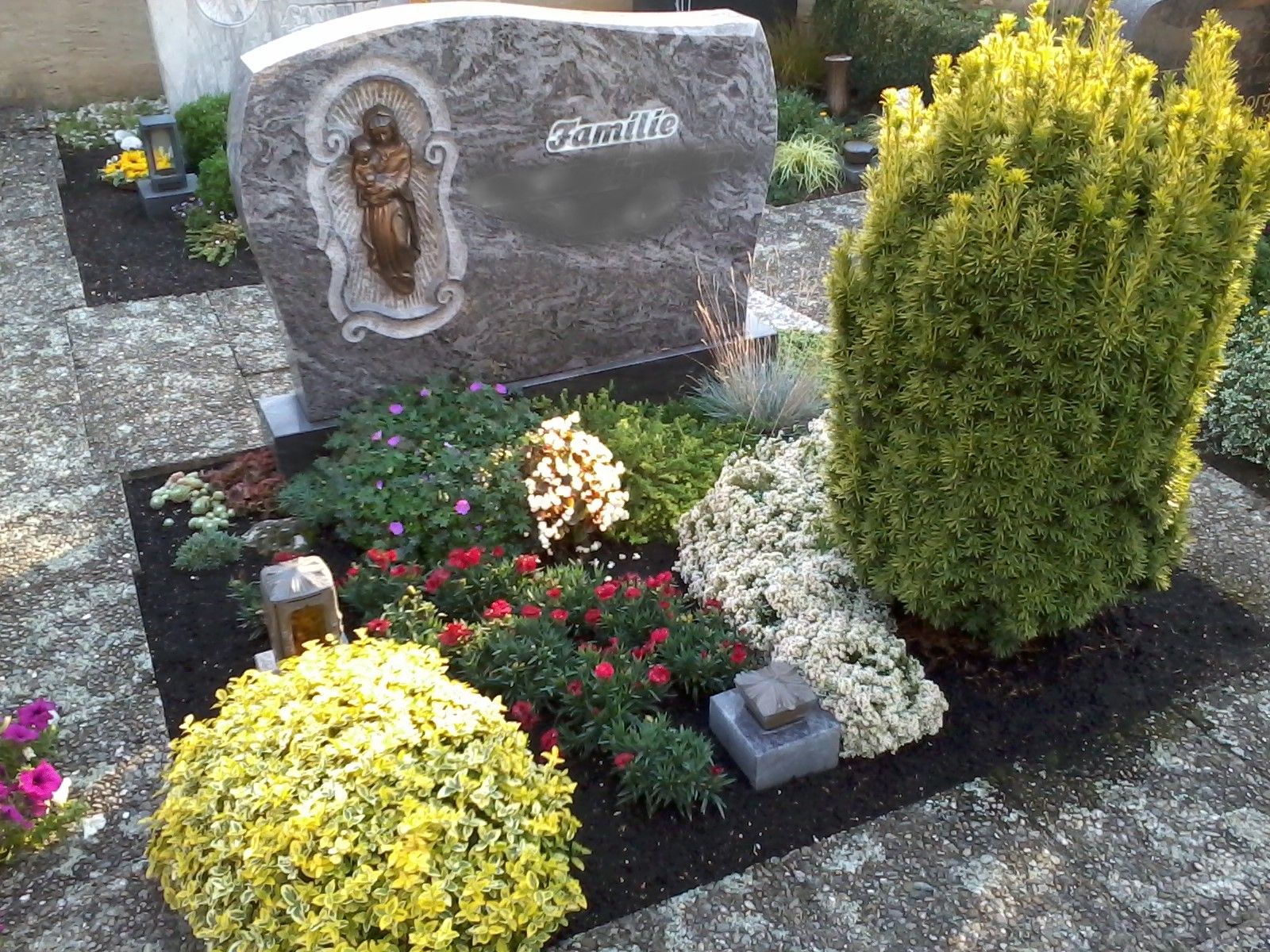 Grabstein mit einer religiösen Figur, umgeben von bunten Blumen und Grün.