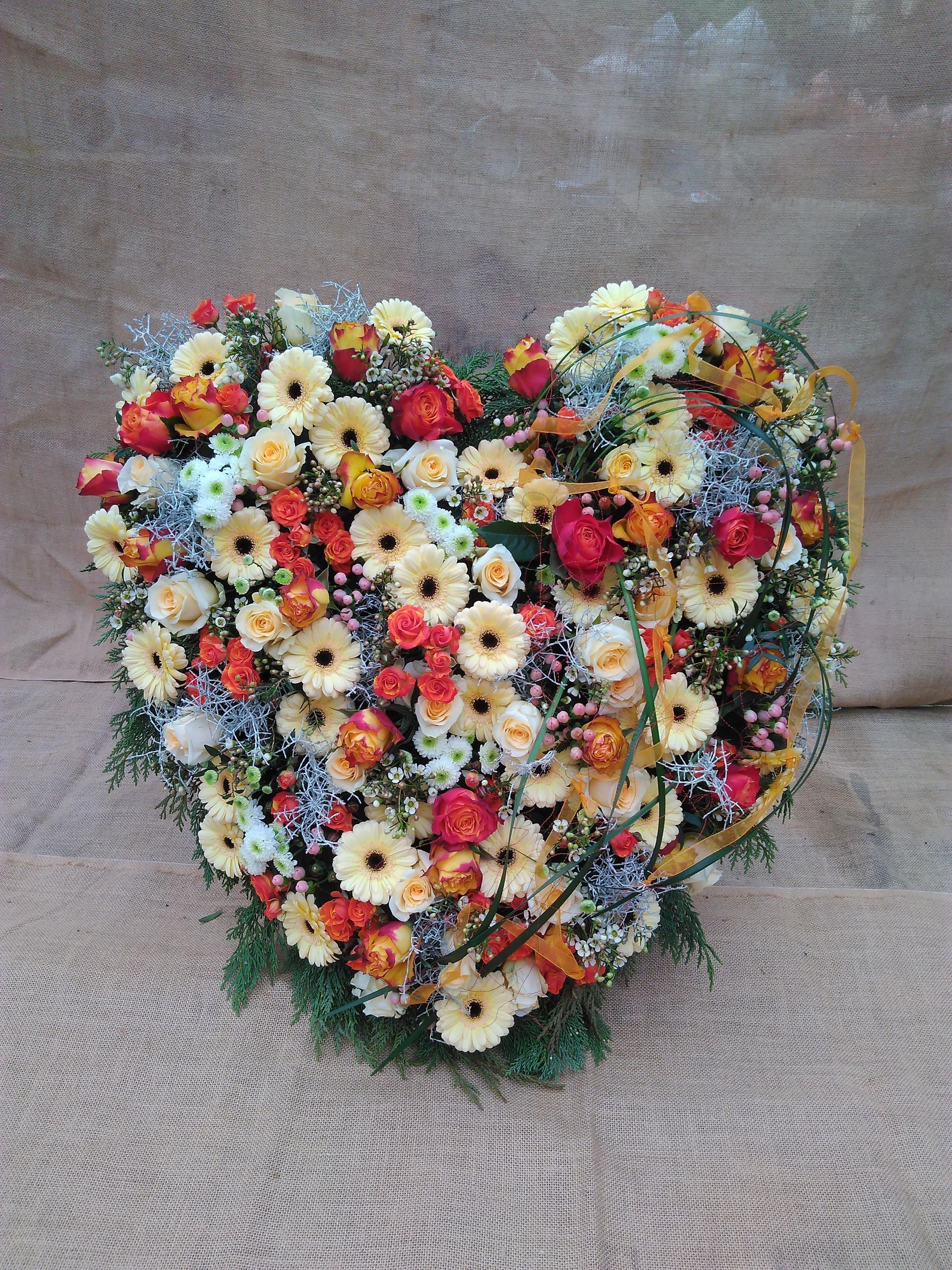 Herzförmiges Blumenarrangement; cremefarbene und orangefarbene Blumen, Grünpflanzen, auf braunem Hintergrund.