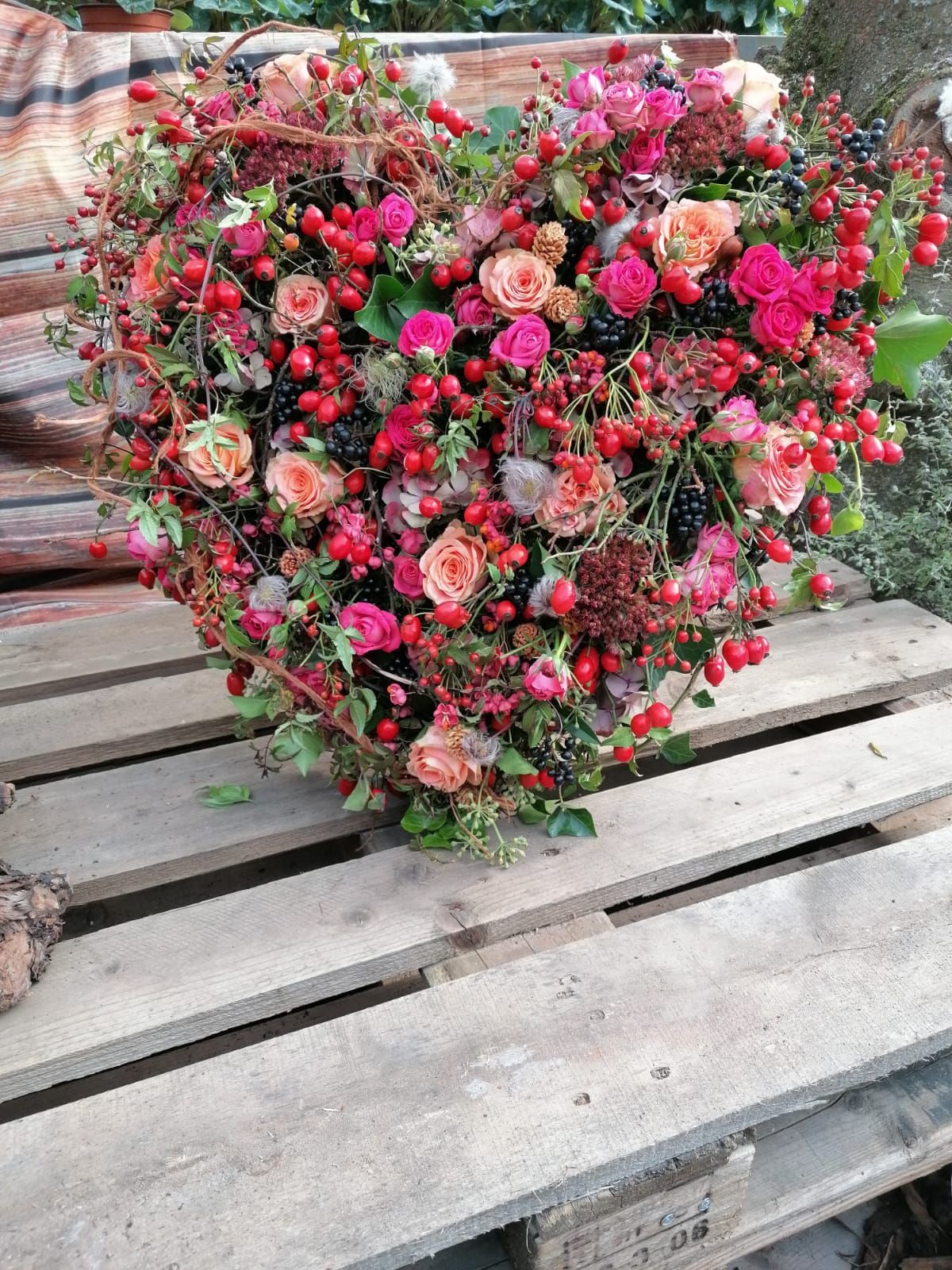 Herzförmiges Blumenarrangement aus Rosen und Beeren auf einer Holzpalette.