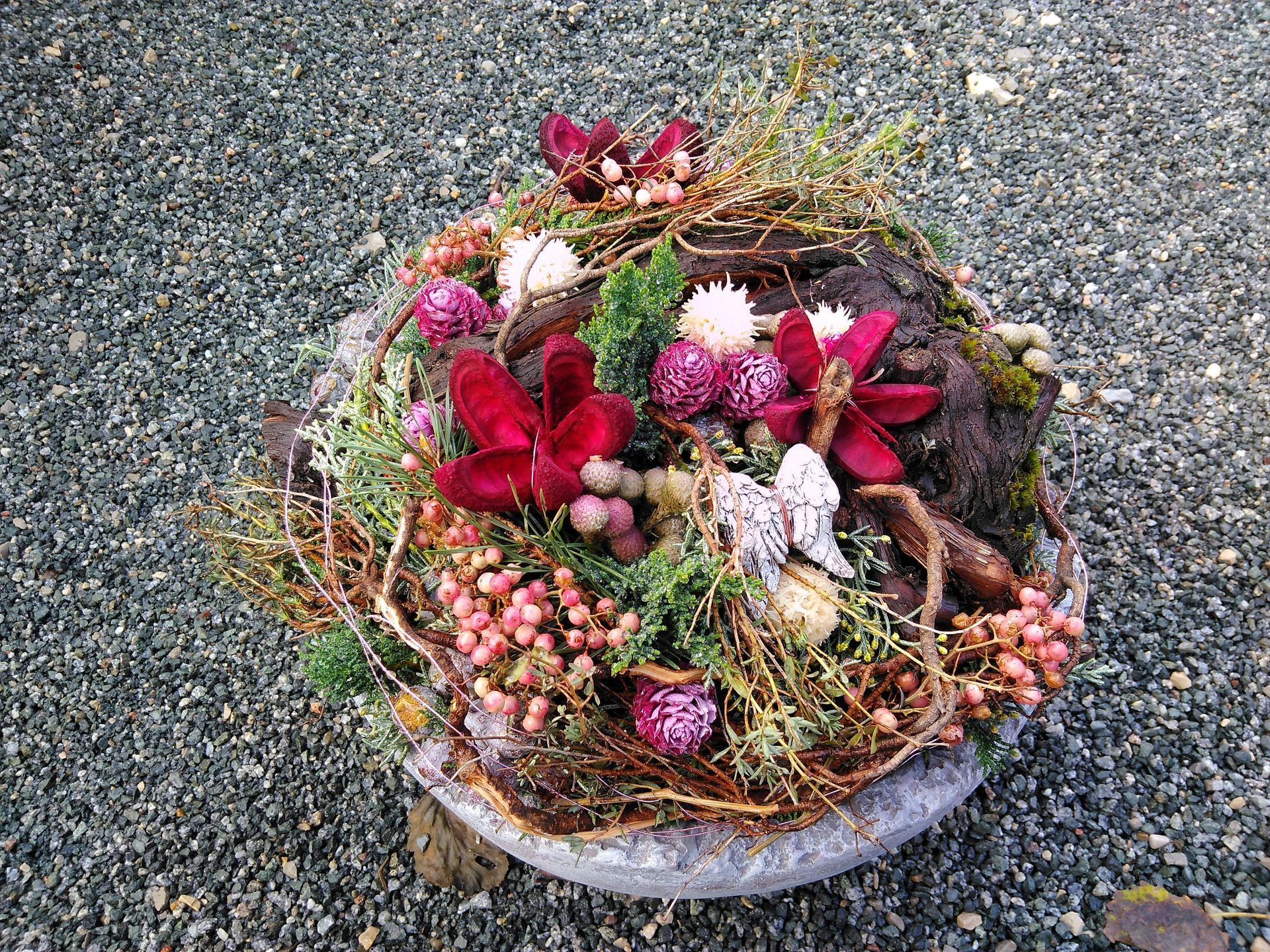 Nahaufnahme eines Blumenarrangements mit dunkelroten und rosa Blüten, eingebettet in einen Steinbehälter vor einem Kieshintergrund.