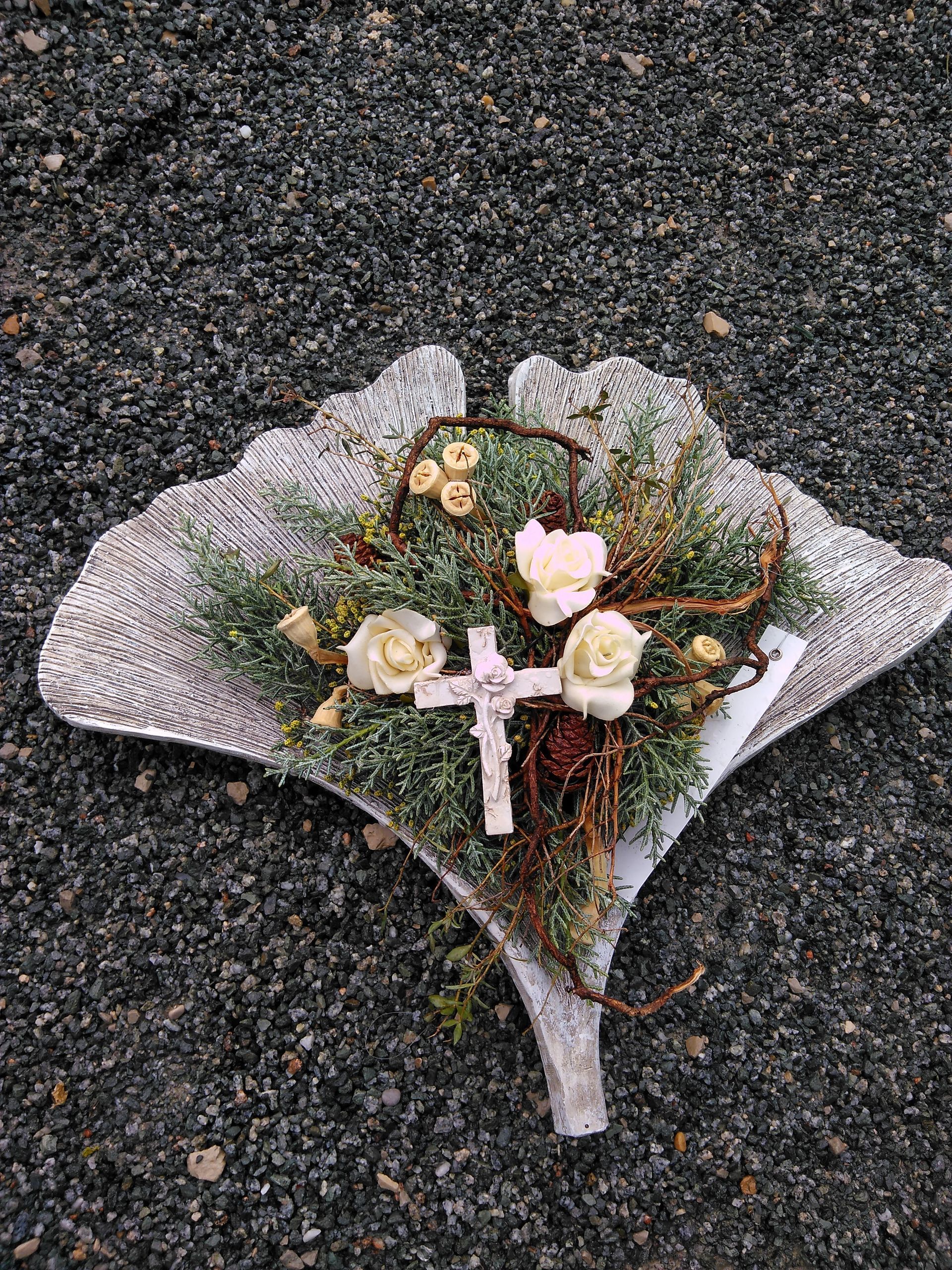 Ein Blumenarrangement in Form eines Blattes mit einem Kreuz, weißen Blüten und grünem Laub auf einer dunkelgrauen Kiesoberfläche.