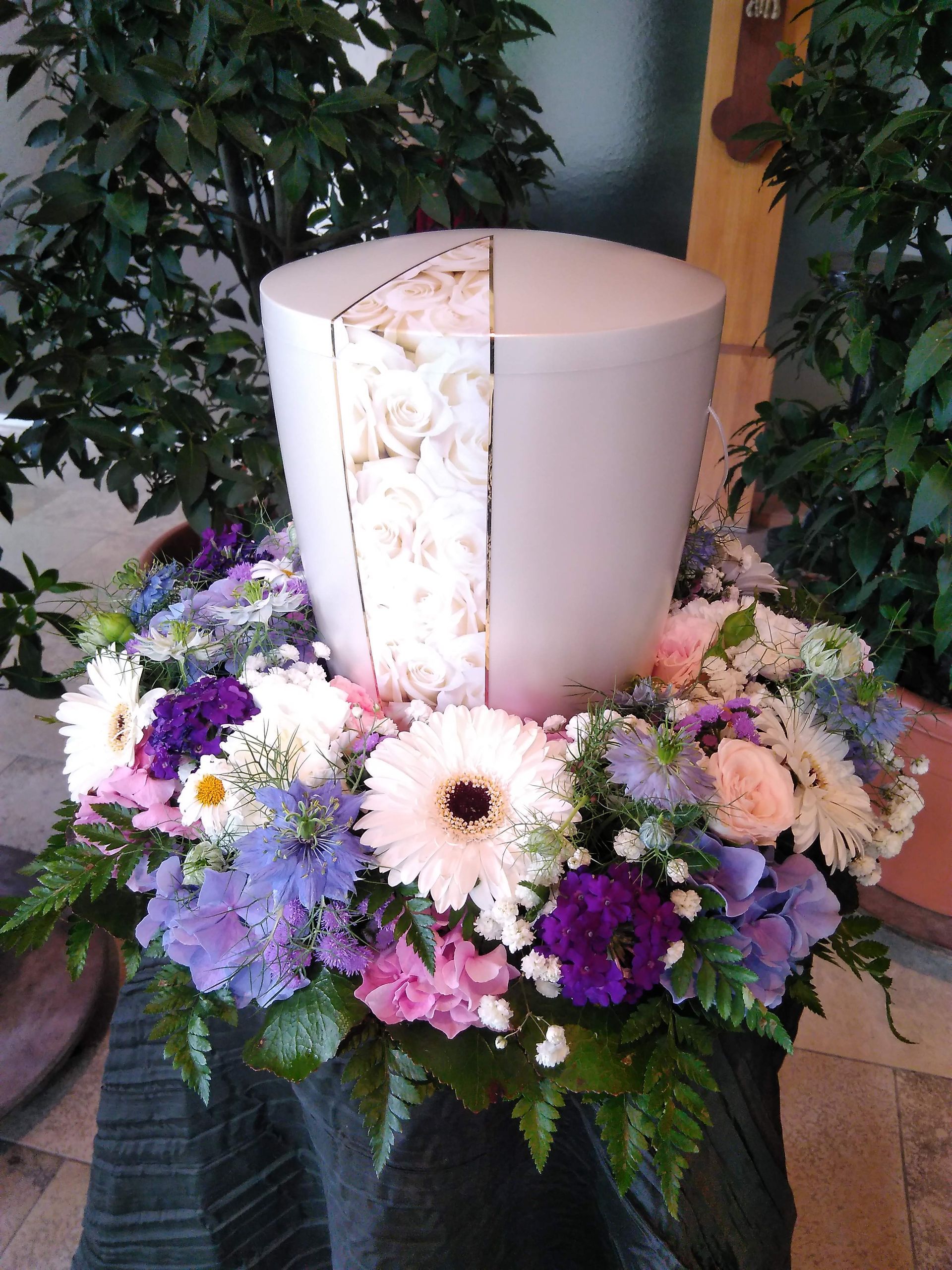 Urne umgeben von einem Blumenarrangement in Lila- und Weißtönen.