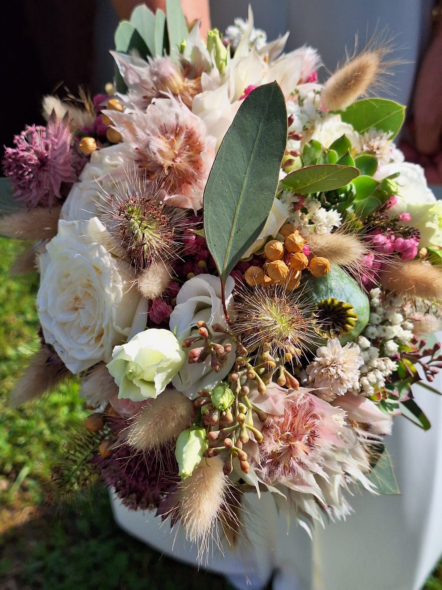 Nahaufnahme eines Brautstraußes mit weißen Rosen, malvenfarbenen Blumen, getrocknetem Weizen, grünem Laub und Akzenten, der von einer Person gehalten wird.