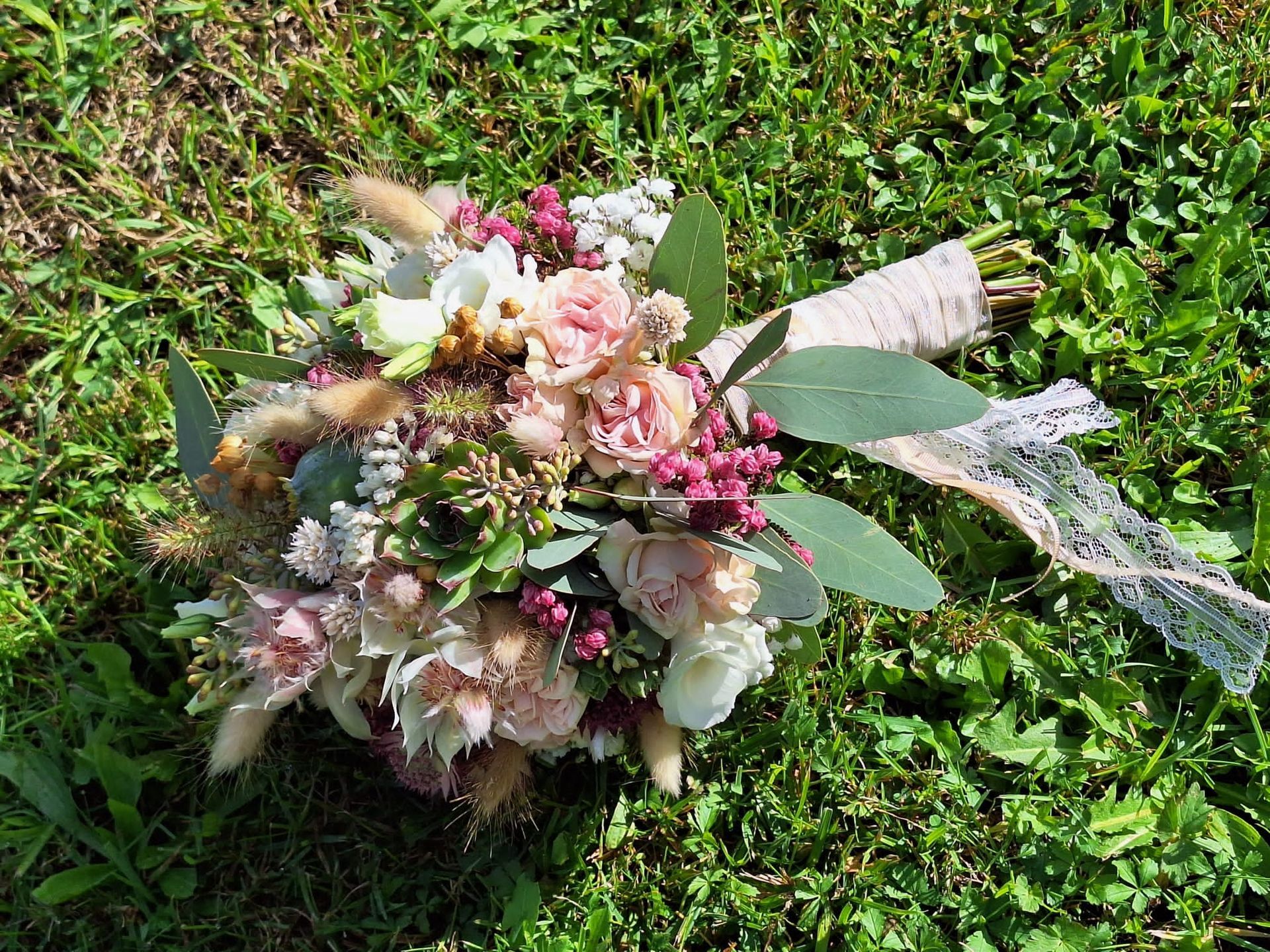 Strauß rosa und weißer Rosen mit grünem Laub, mit Spitzenband zusammengebunden, auf grünem Gras liegend.