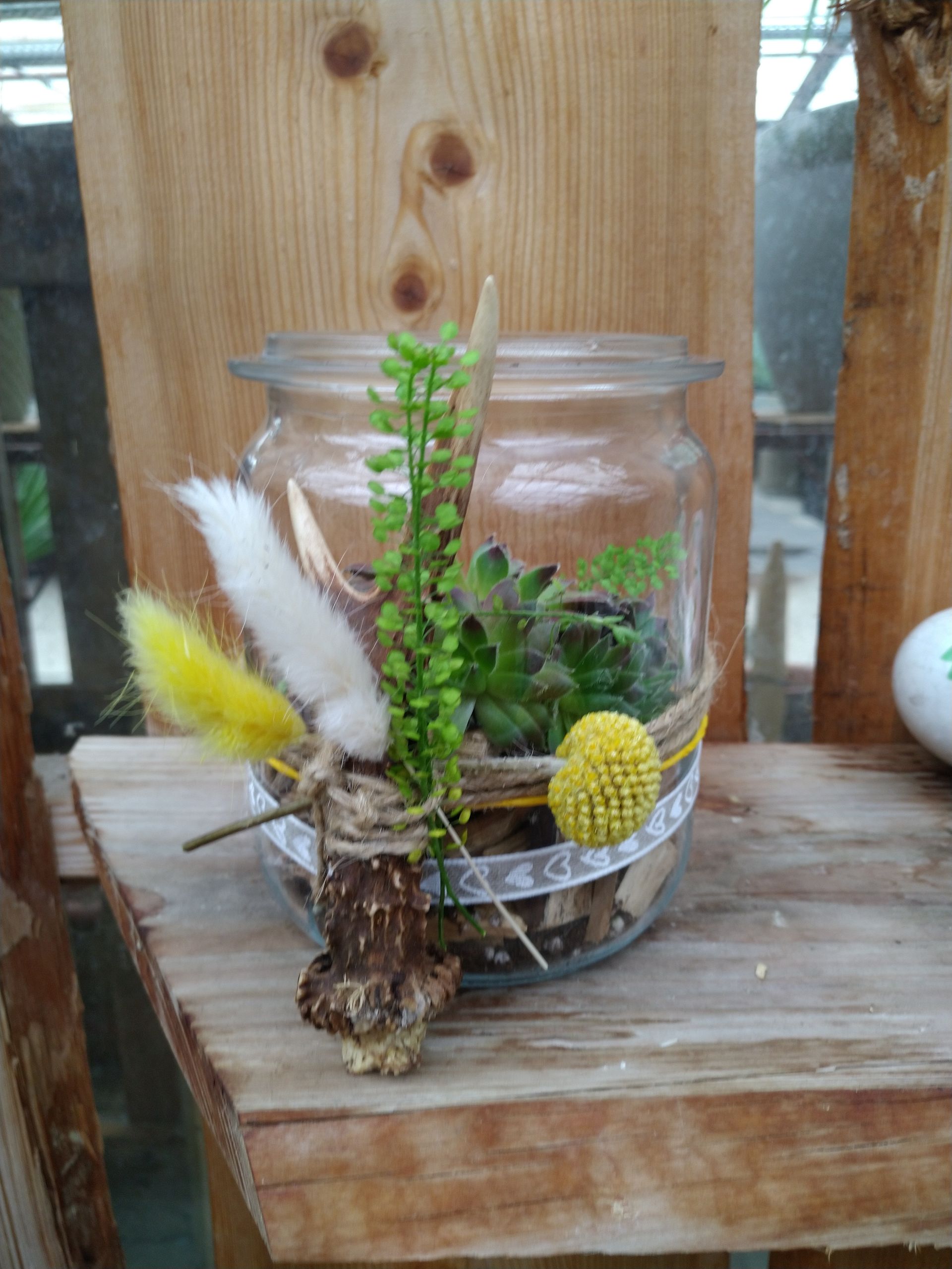 Terrarium in einem Glasgefäß mit getrockneten Blumen und Kunstpflanzen auf einem Holzregal.