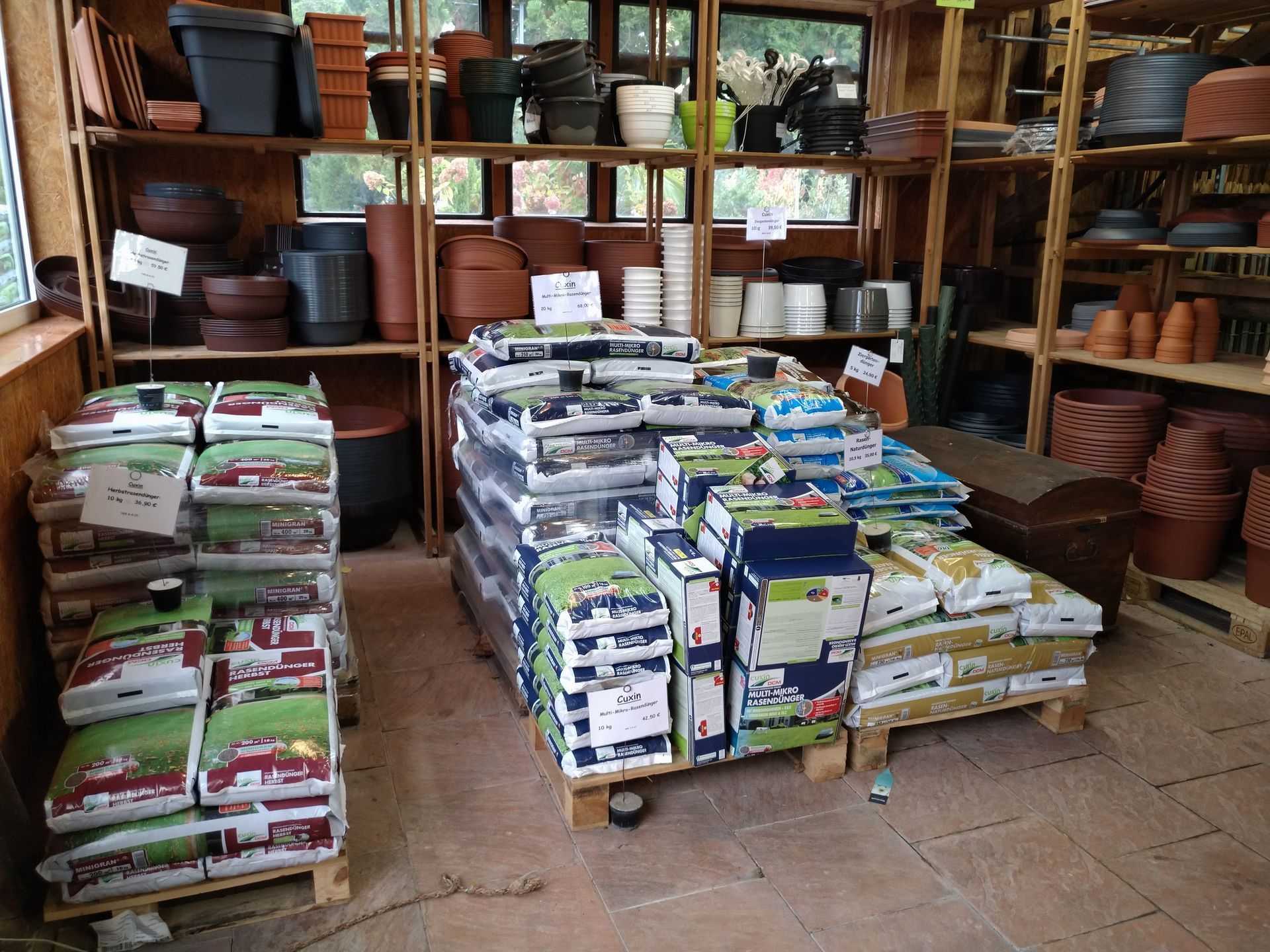 Regale mit Gartenbedarf in einem Geschäft: Auf Paletten gestapelte Erdsäcke, Töpfe und Behälter in einem Holzschuppen.