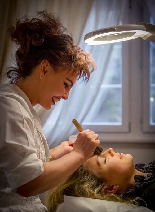 Une esthéticienne maquille une cliente sous une lampe frontale éclairée par une lampe frontale près d'une fenêtre.