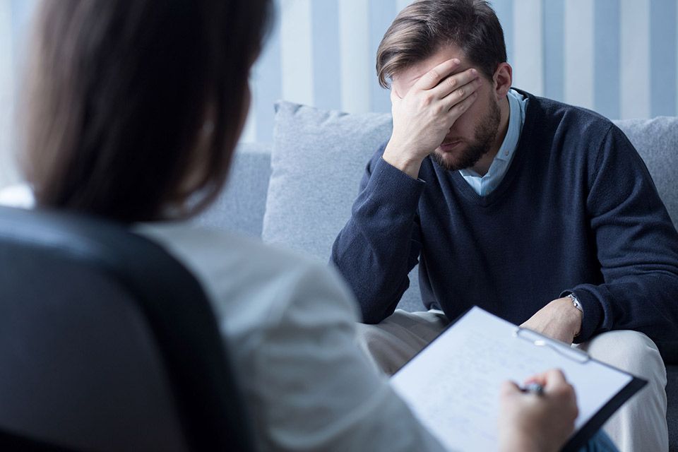 Un hombre está sentado en un sofá hablando con una mujer que sostiene un portapapeles.